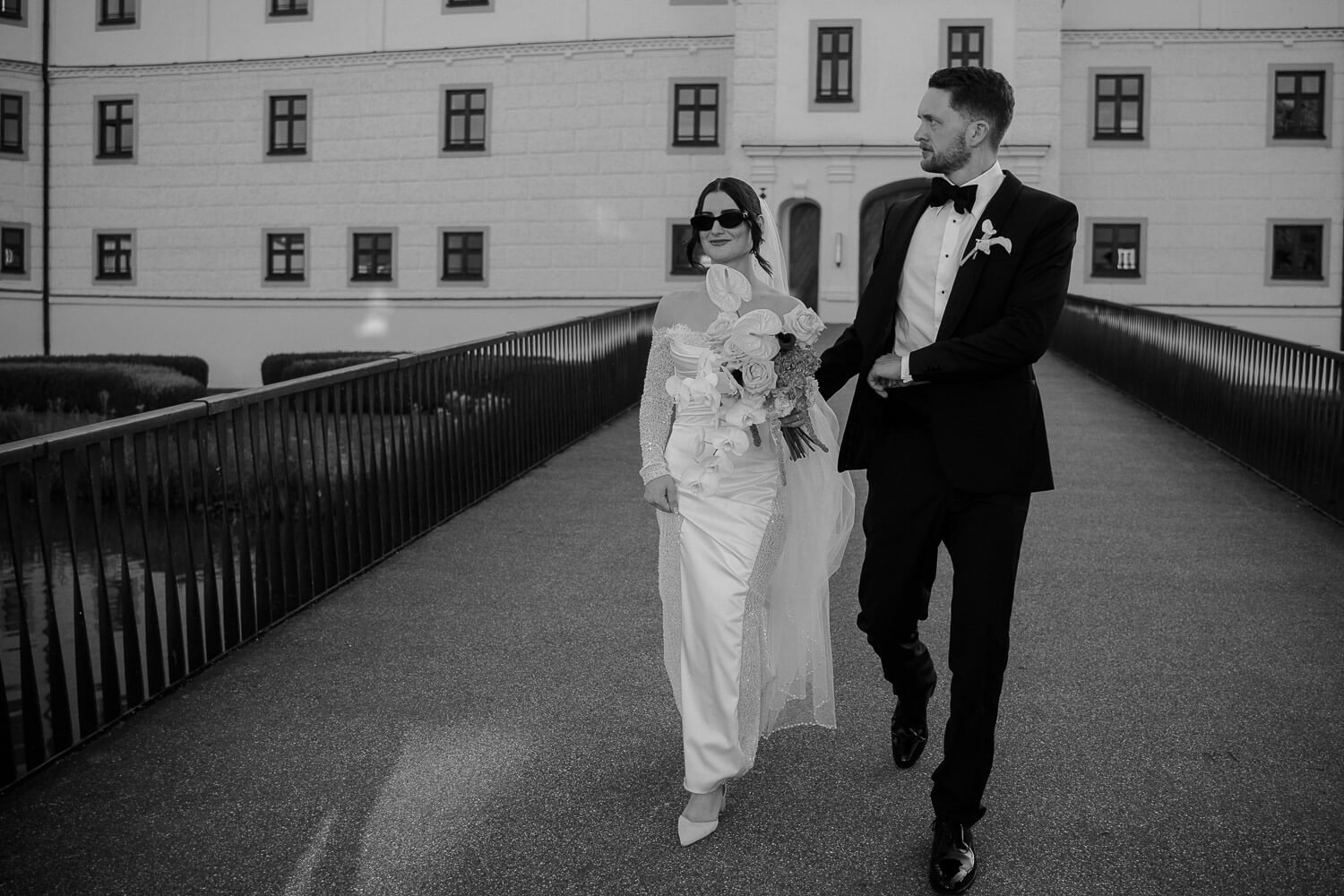 Stilvolles Hochzeitspaar auf der Brücke von Schloss Hohenkammer, Braut mit Sonnenbrille und Blumenstrauß.