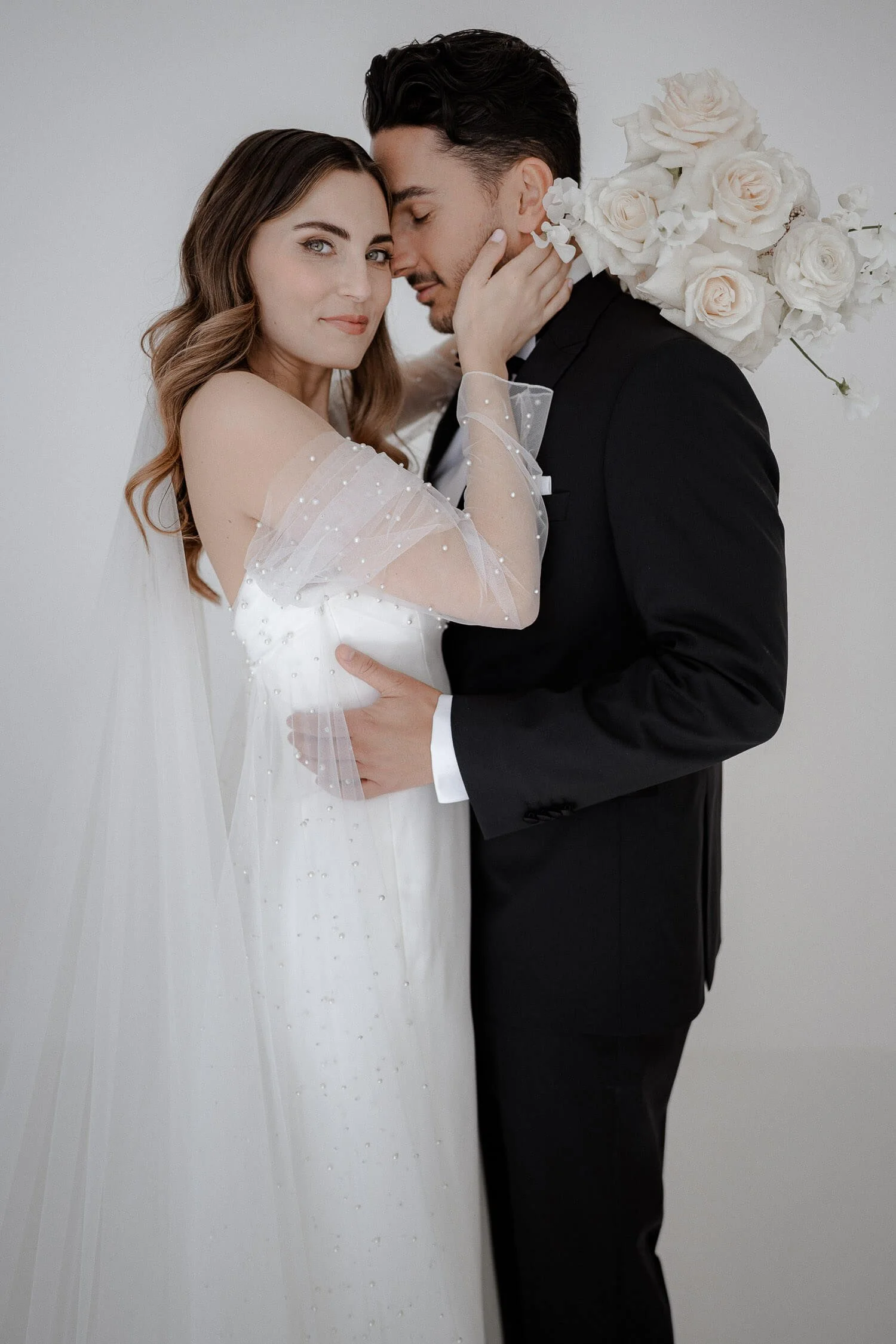 Elegantes Brautpaar innig umarmt vor weißem Hintergrund, fotografiert von einem Hochzeitsfotografen in München.