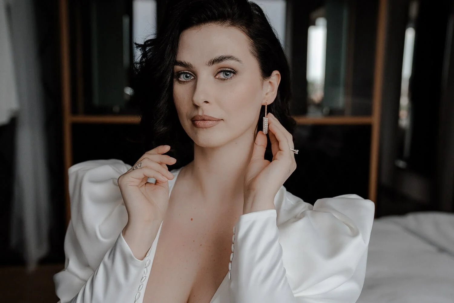 Elegante Braut beim Getting Ready im Hotel Andaz in München – Frau mit dunklen Locken und weißen Ärmeln setzt Perlenohrring ein.