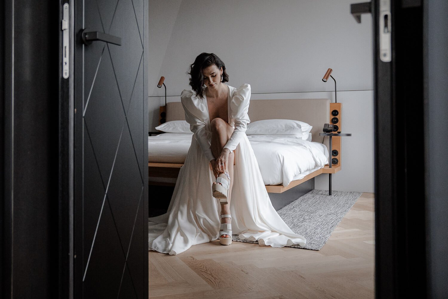 Braut im Andaz Schwabinger Tor München – Elegante Frau im Hochzeitskleid sitzt im Hotelzimmer und zieht ihre Schuhe an.