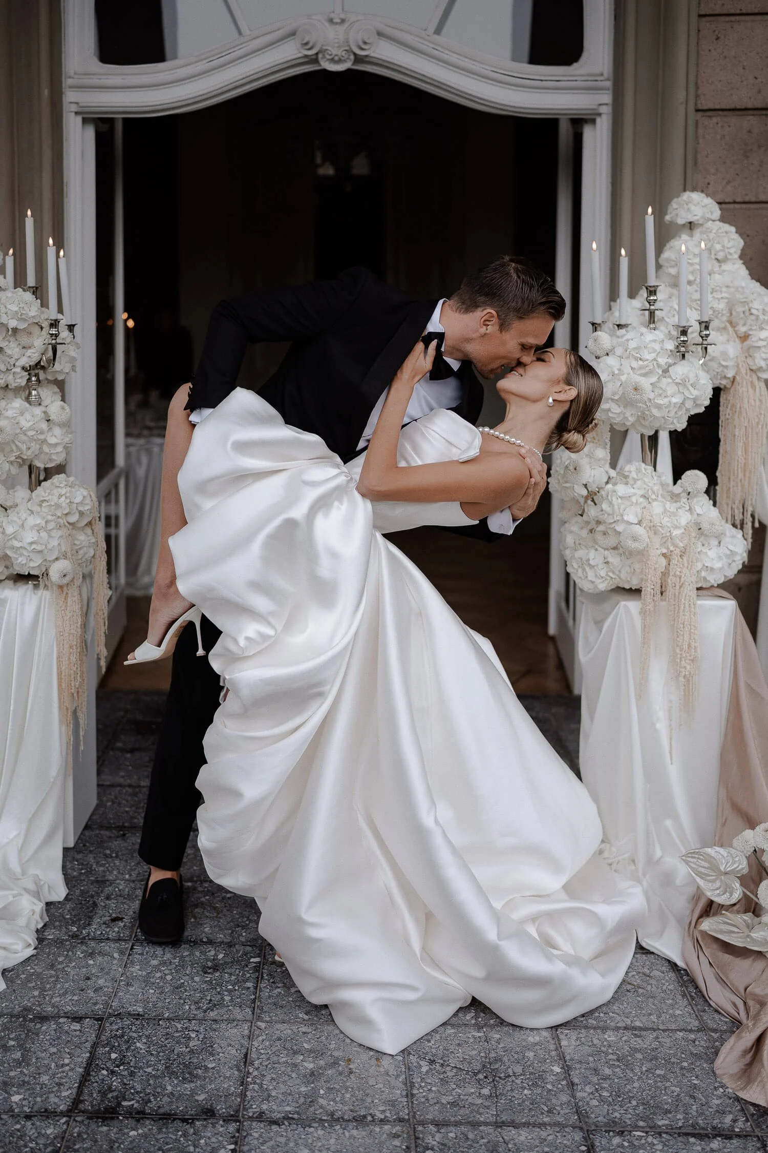 Brautmode München: Brautpaar küsst sich, Braut im Satin-Brautkleid vor Blumen-Deko