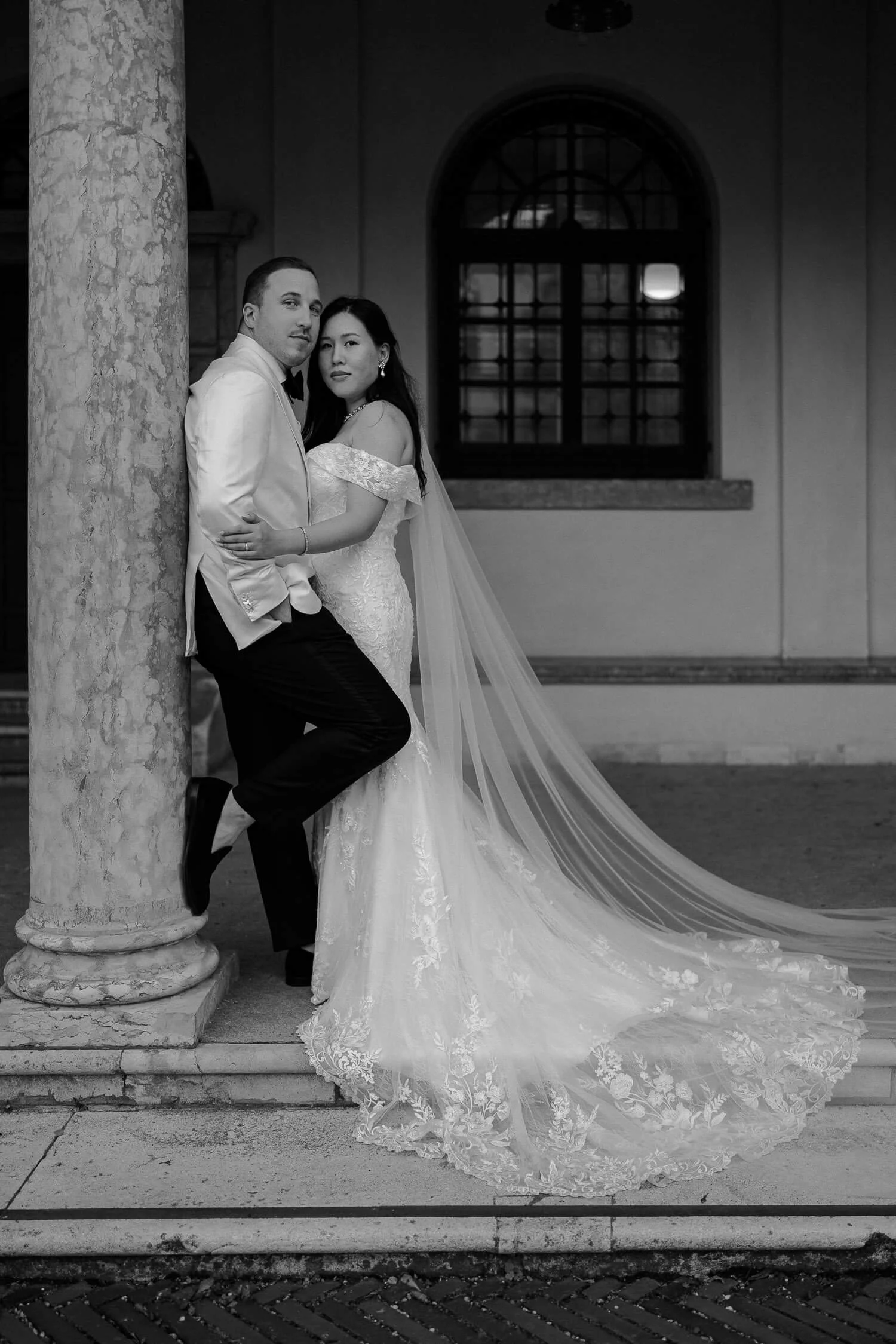 Elegantes Hochzeitspaar in München – Braut mit langem Schleier lehnt an Bräutigam vor historischer Säule.
