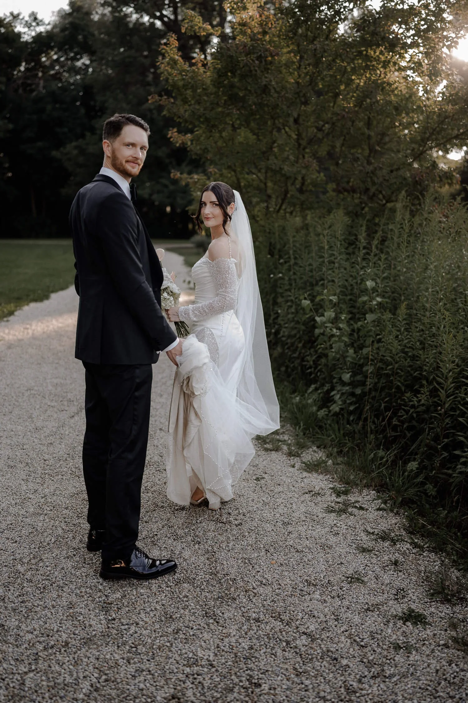 Hochzeitspaar in Schloss Hohenkammer beim Spaziergang im Abendlicht, Bräutigam hält das Kleid der Braut.