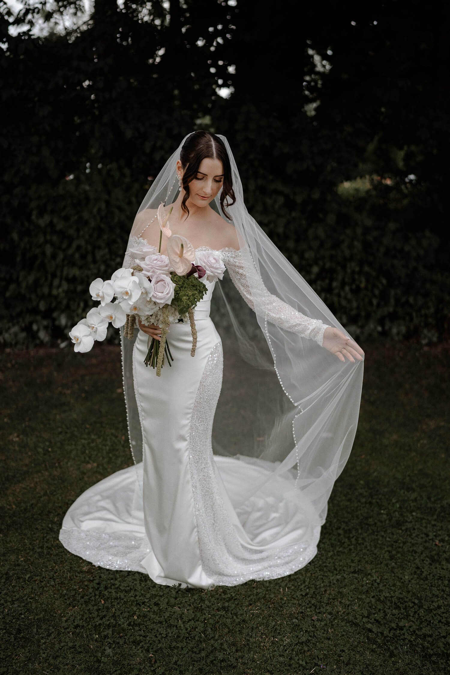 Elegante Braut in Schloss Hohenkammer bei München mit langem Schleier und edlem Brautstrauß vor dunkler Naturkulisse.