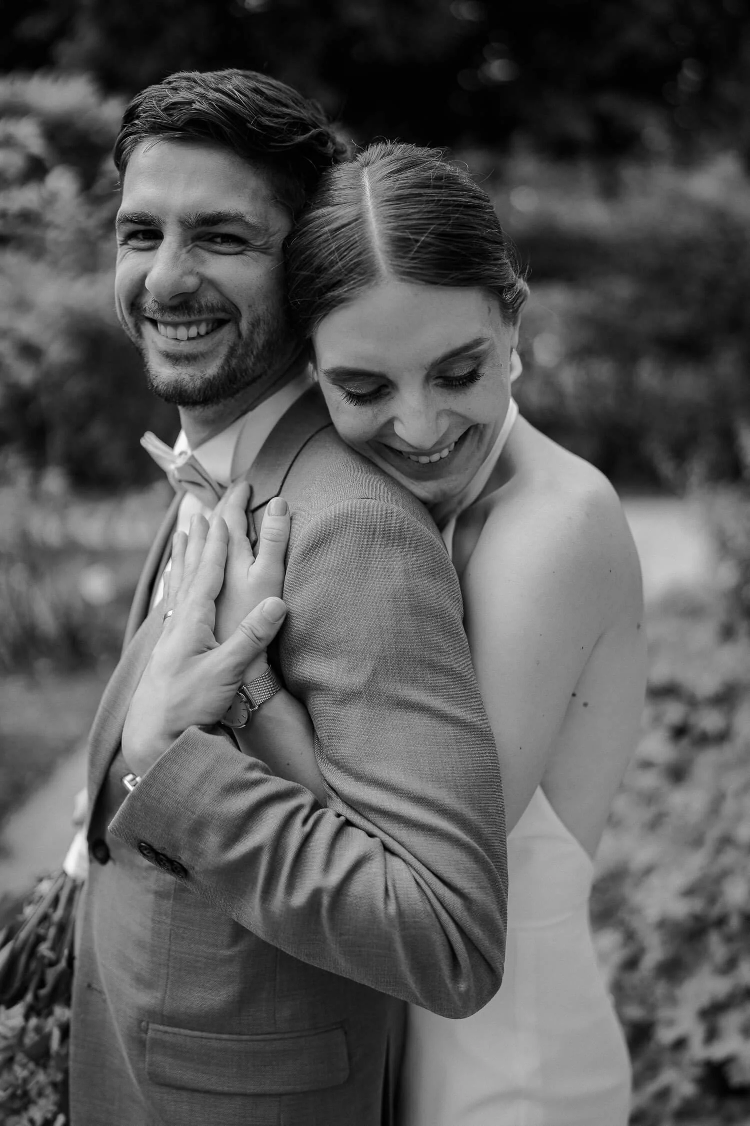 Intimes Close-up, Braut umarmt Bräutigam von hinten/seitlich, beide lächeln – brautpaar umarmung close up sw emotional wedding portrait