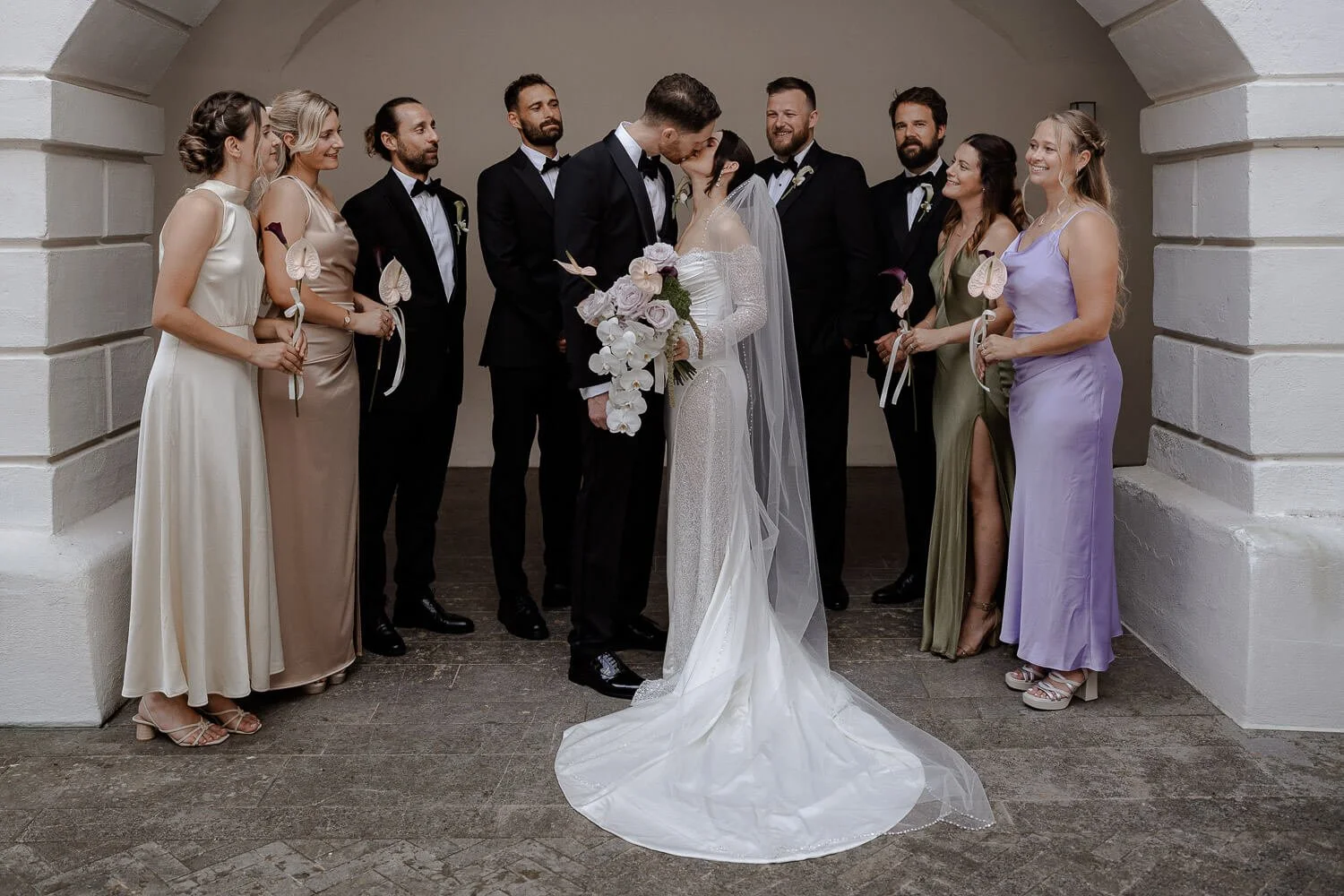 Brautpaar küsst sich mit Trauzeugen im Hintergrund.