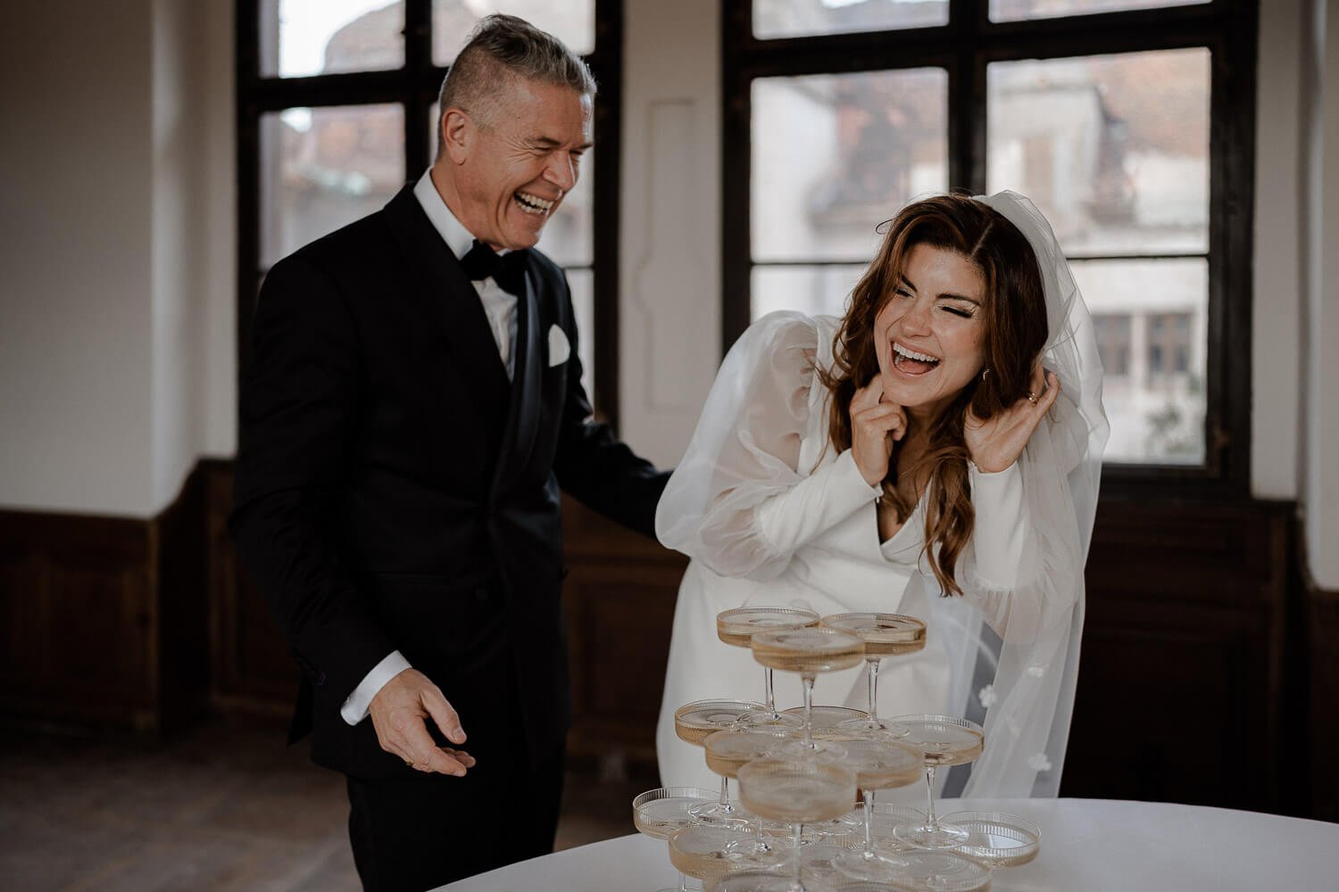 Lachende Braut mit Bräutigam vor Champagnerturm – ausgelassene Hochzeitsfeier aufgenommen von Hochzeitsfotografin aus München.