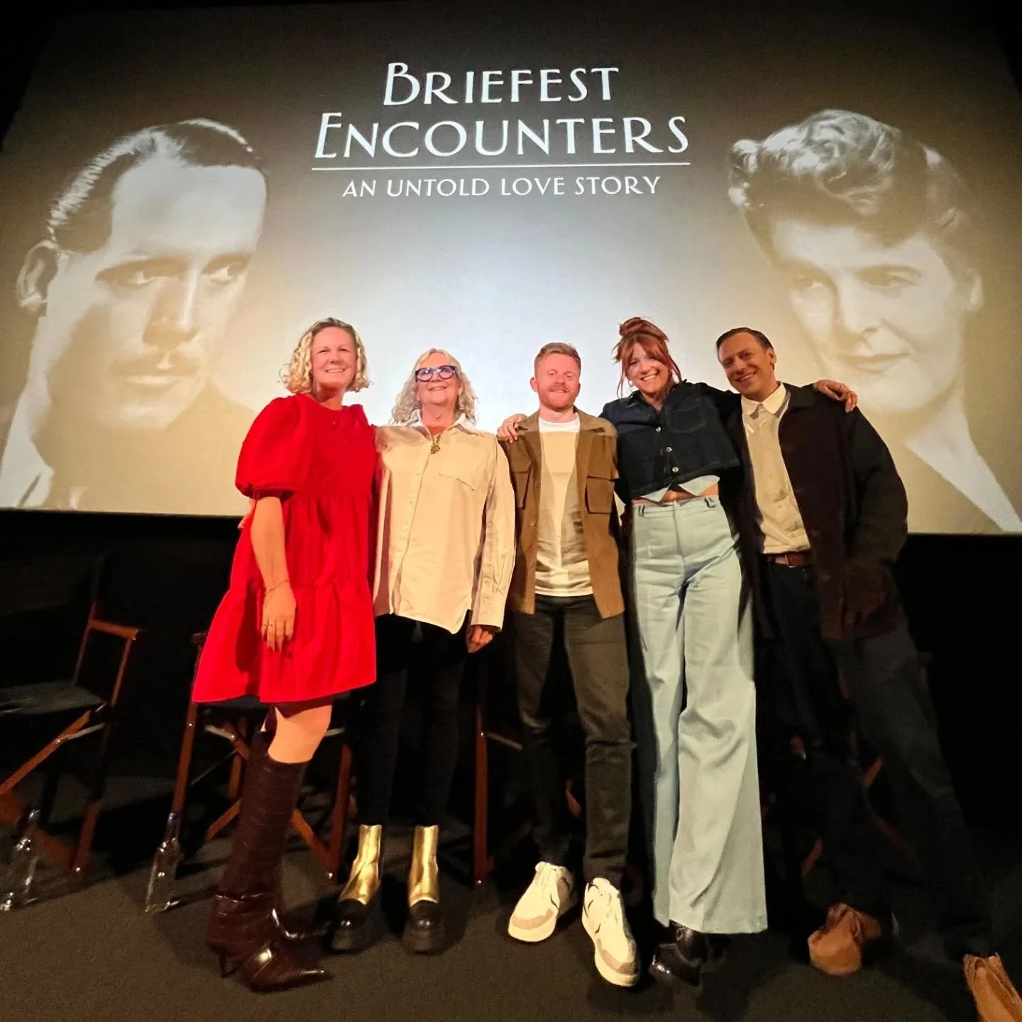 Group of five people standing in front of a movie poster for "Brief Encounters: An Untold Love Story" with black-and-white images of a man and woman. The group includes three women and two men, all smiling and posing for the photo, with some individuals wearing casual or semi-casual clothing.
