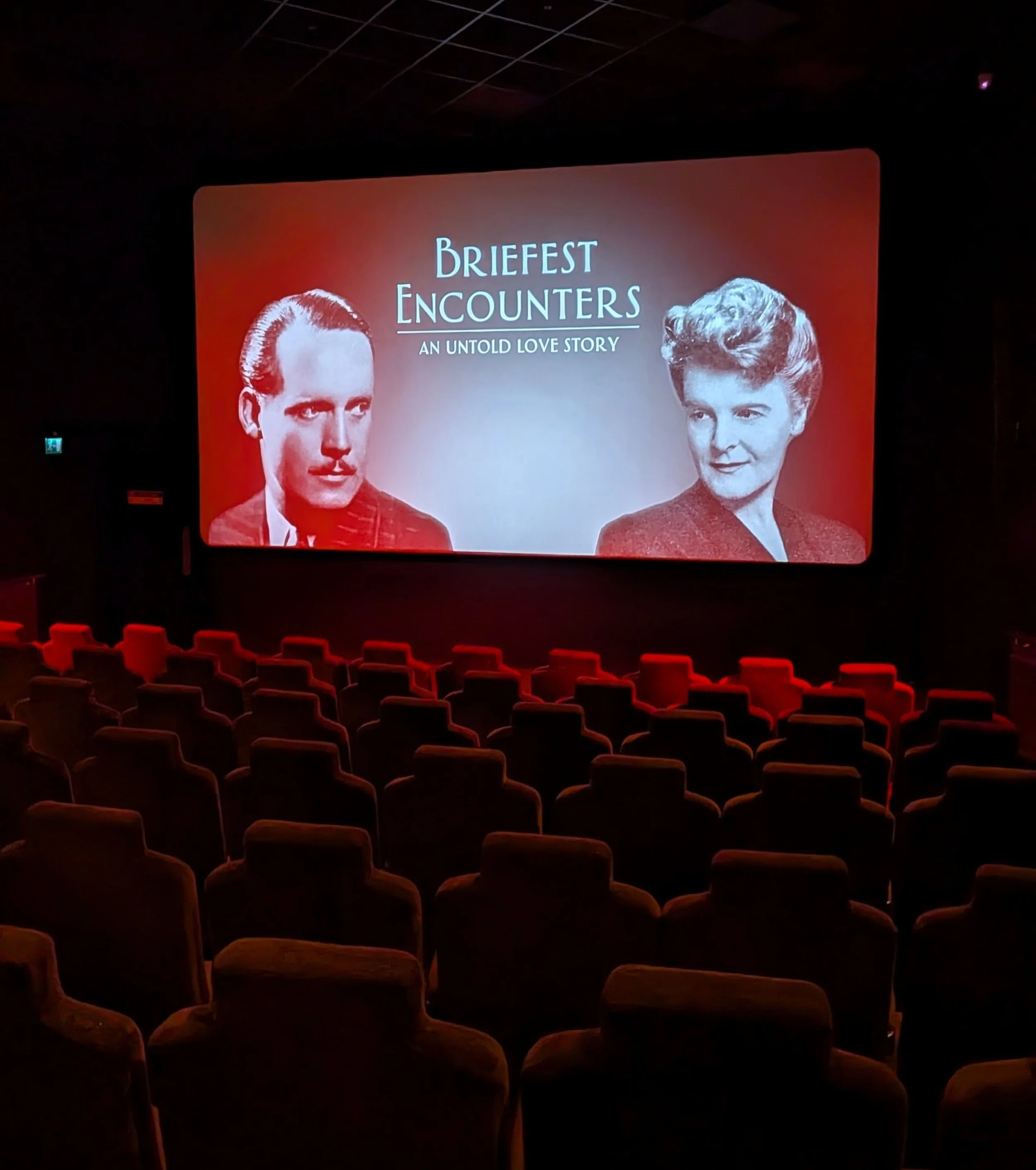 Empty theater or auditorium with chairs facing a large screen displaying an event titled 'Briefest Encounters: An Untold Love Story,' featuring black and white photos of a man and a woman.
