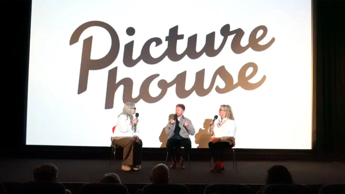 Three people sitting on stage in front of a large screen that reads 'Picture House,' holding microphones and engaging in a discussion.