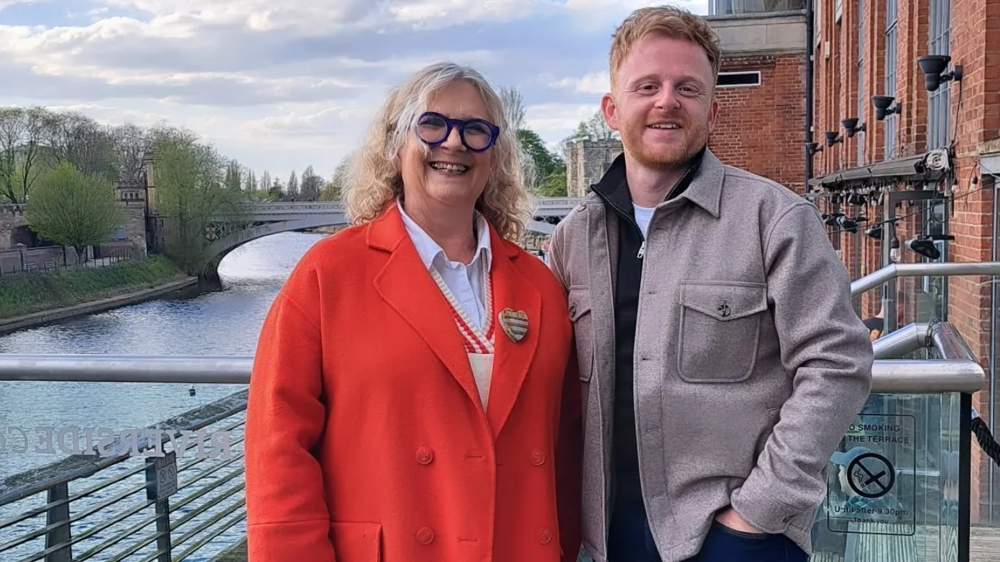 A smiling woman with blond hair, glasses, wearing a red coat, and a man with red hair, wearing a gray jacket, standing outdoors on a bridge over a river with a brick building behind them.