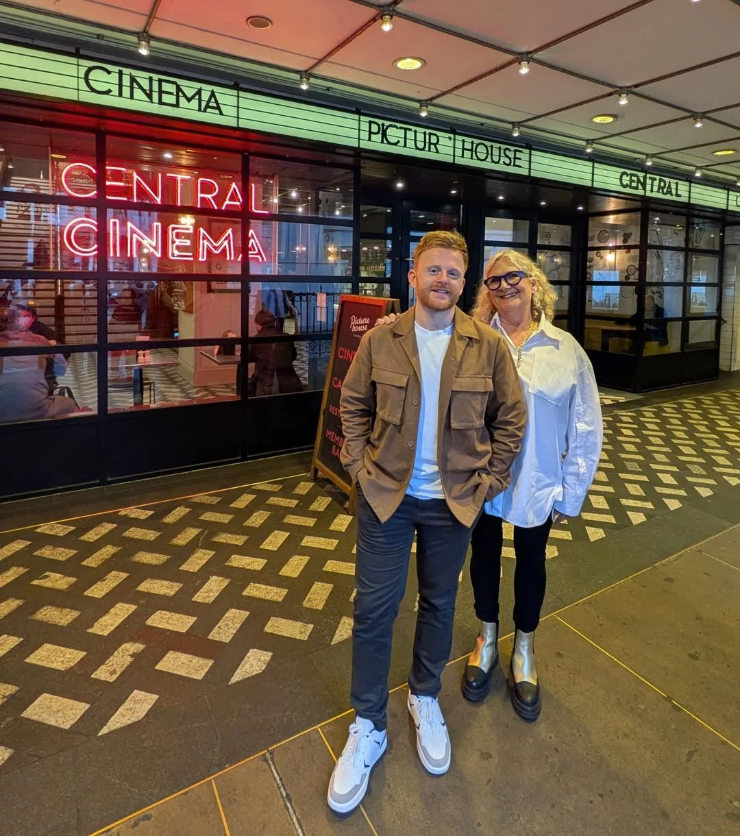 A man and woman standing outside a theater with a bright neon sign that reads 'Central Cinema'. The theater has a glass exterior with visible interior seating and people inside. The man is wearing a tan jacket, white shirt, dark pants, and sneakers, while the woman is wearing a white jacket, black pants, chunky shoes, and glasses. Both are smiling and posing for the photo.