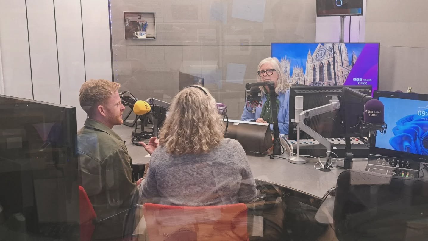 Three people inside a radio broadcasting studio, two women and one man, with recording equipment, microphones, and monitors showing the BBC Radio York logo.