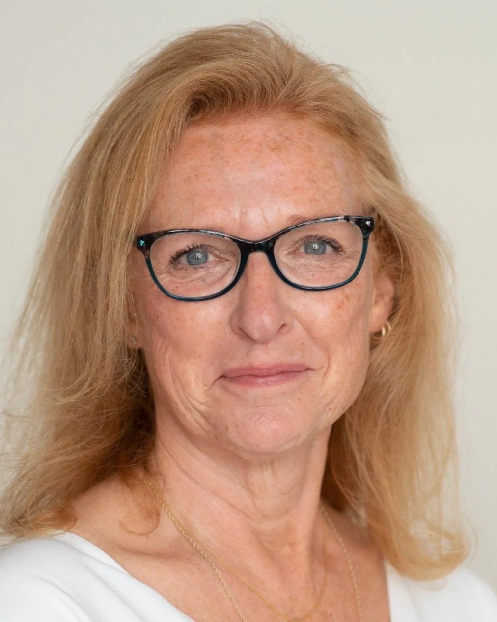 Close-up portrait of a mature woman with red hair, glasses, and a slight smile, wearing a white top and a gold necklace.