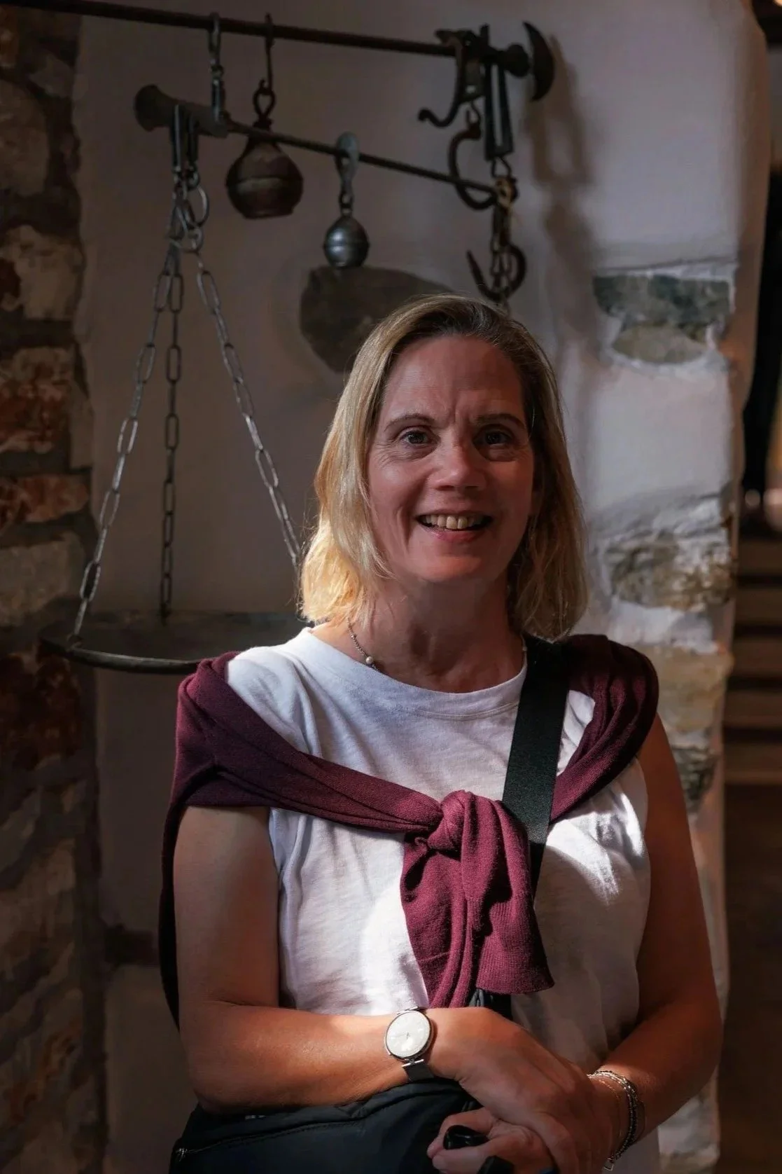 A woman with light-colored shoulder-length hair, smiling, wearing a white sleeveless top with a maroon sweater tied over her shoulders, standing indoors near a stone wall and a vintage balance scale with hanging weights.