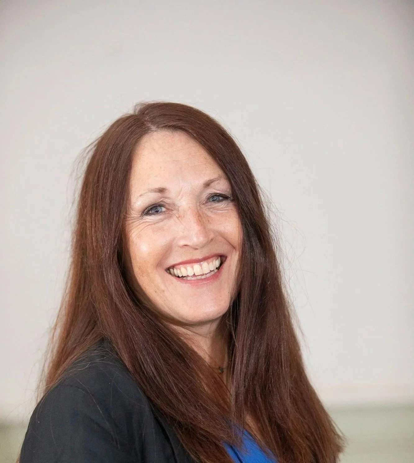 Portrait of a smiling woman with long brown hair, wearing a black blazer and blue top, standing against a plain light background.