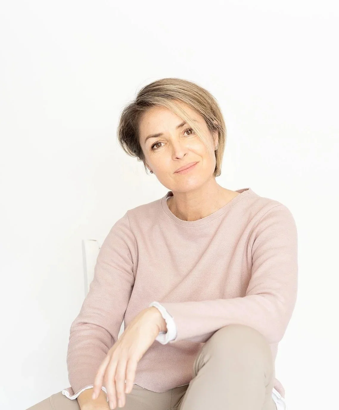 A woman with short light brown hair and wearing a light pink sweater sitting against plain white background.