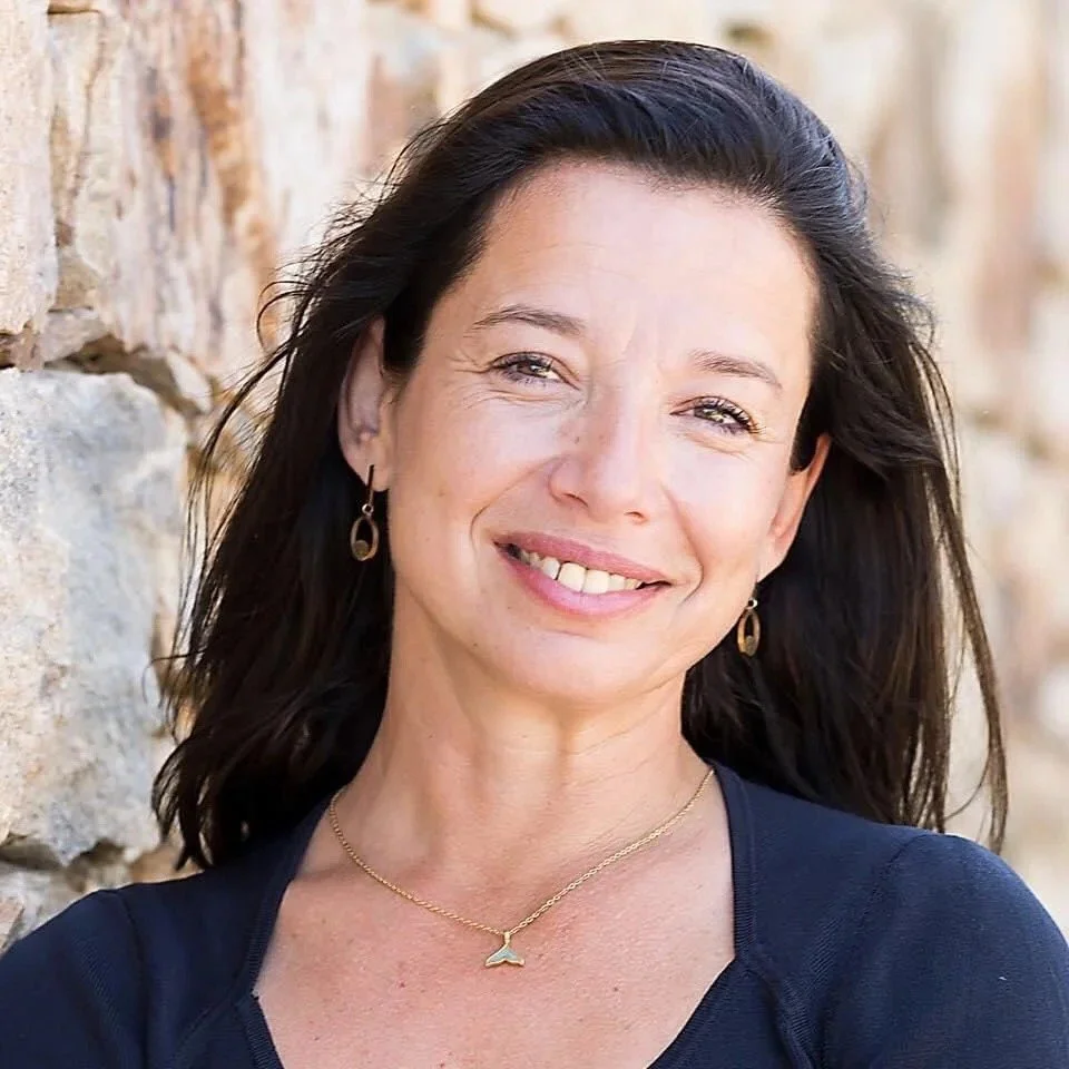 A smiling woman with dark hair, wearing gold earrings and a dolphin necklace, standing near a stone wall.