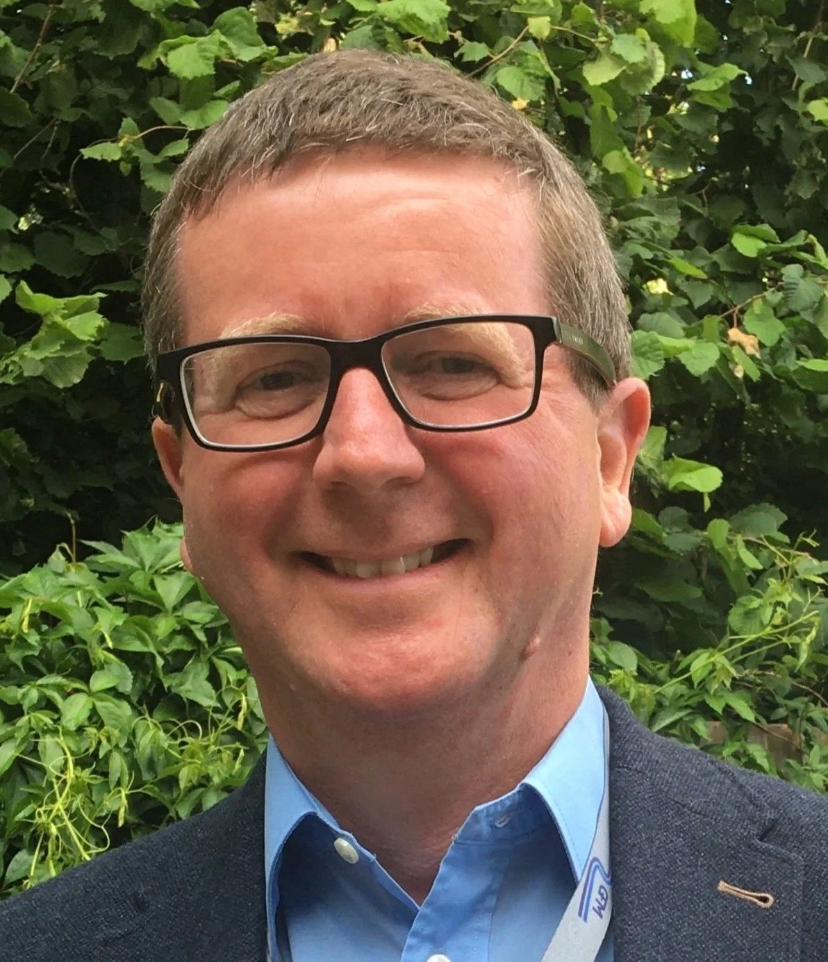 A smiling man with short brown hair, wearing black glasses, a blue shirt, and a dark blazer, standing in front of green leafy bushes.