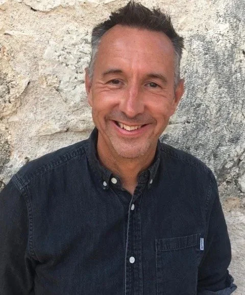 A smiling man with short dark hair and a stubble beard, wearing a dark button-up shirt, standing against a textured beige stone wall.