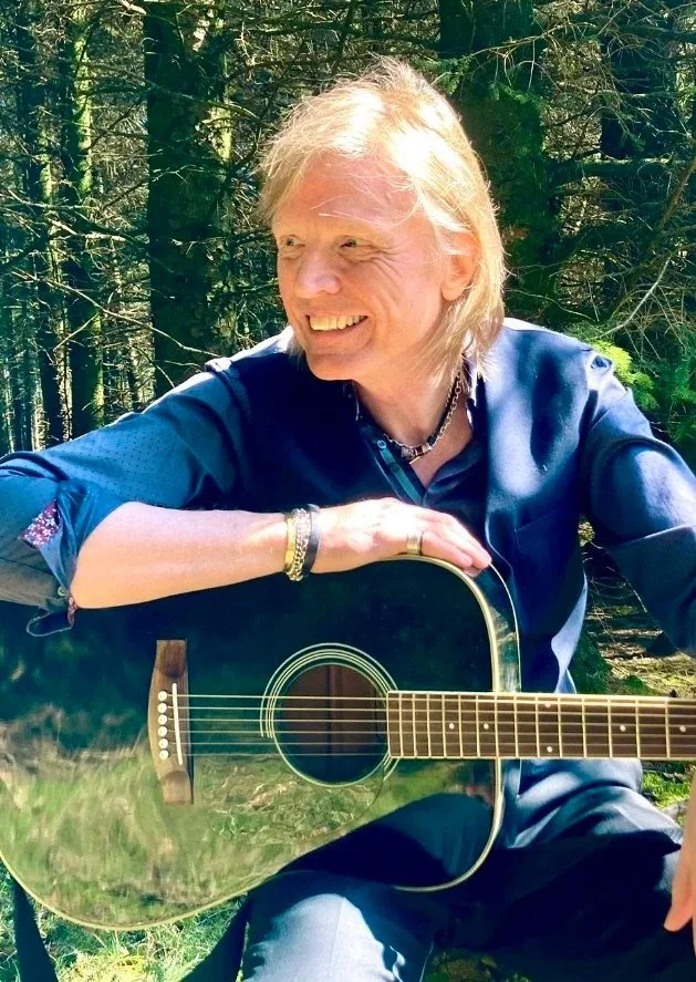 An older woman with blonde hair smiling outdoors in a forest, holding an acoustic guitar.