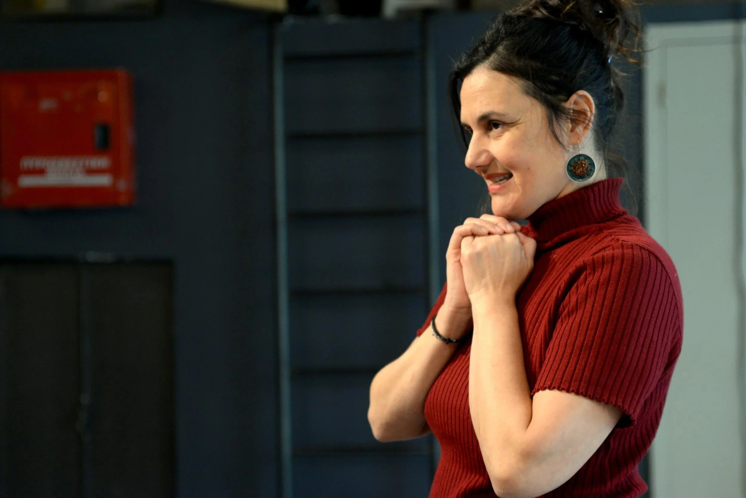 A woman with dark hair tied up, wearing a red turtleneck sweater and large circular earrings, holds her hands together near her chin and appears excited or joyful.