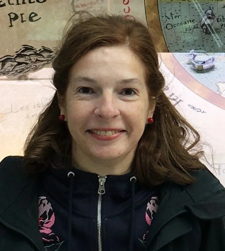 A woman with shoulder-length brown hair, smiling, wearing a black jacket with a zipper and red earrings, standing in front of a colorful wall with illustrations and text.