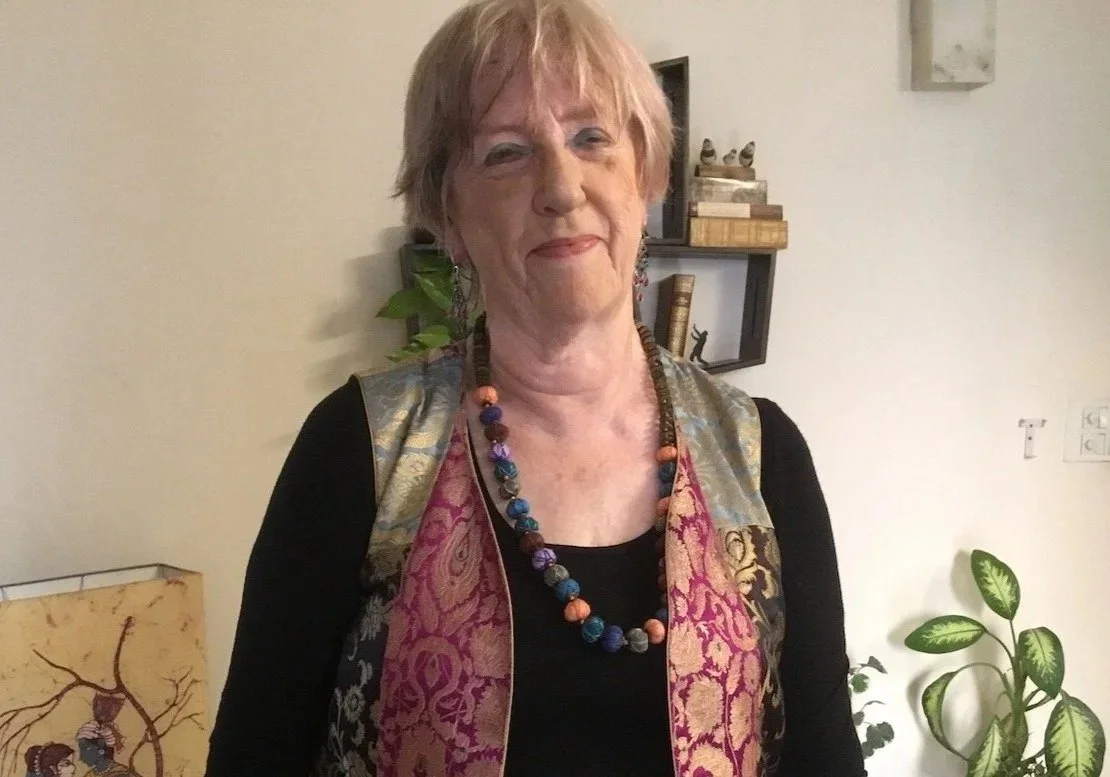 An older woman with short, light-colored hair wearing a colorful, patterned vest and a beaded necklace, standing indoors with books and plants in the background.