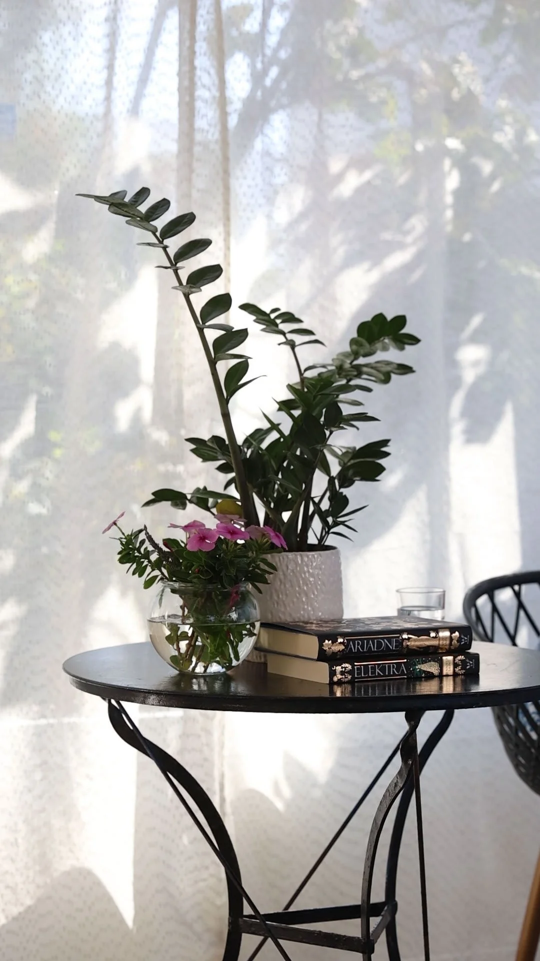 A round black metal table with a white textured plant pot, a small glass vase with pink flowers, two hardcover books, a drinking glass, and a black chair in front of a window with sheer curtains, sunlight casting shadows.