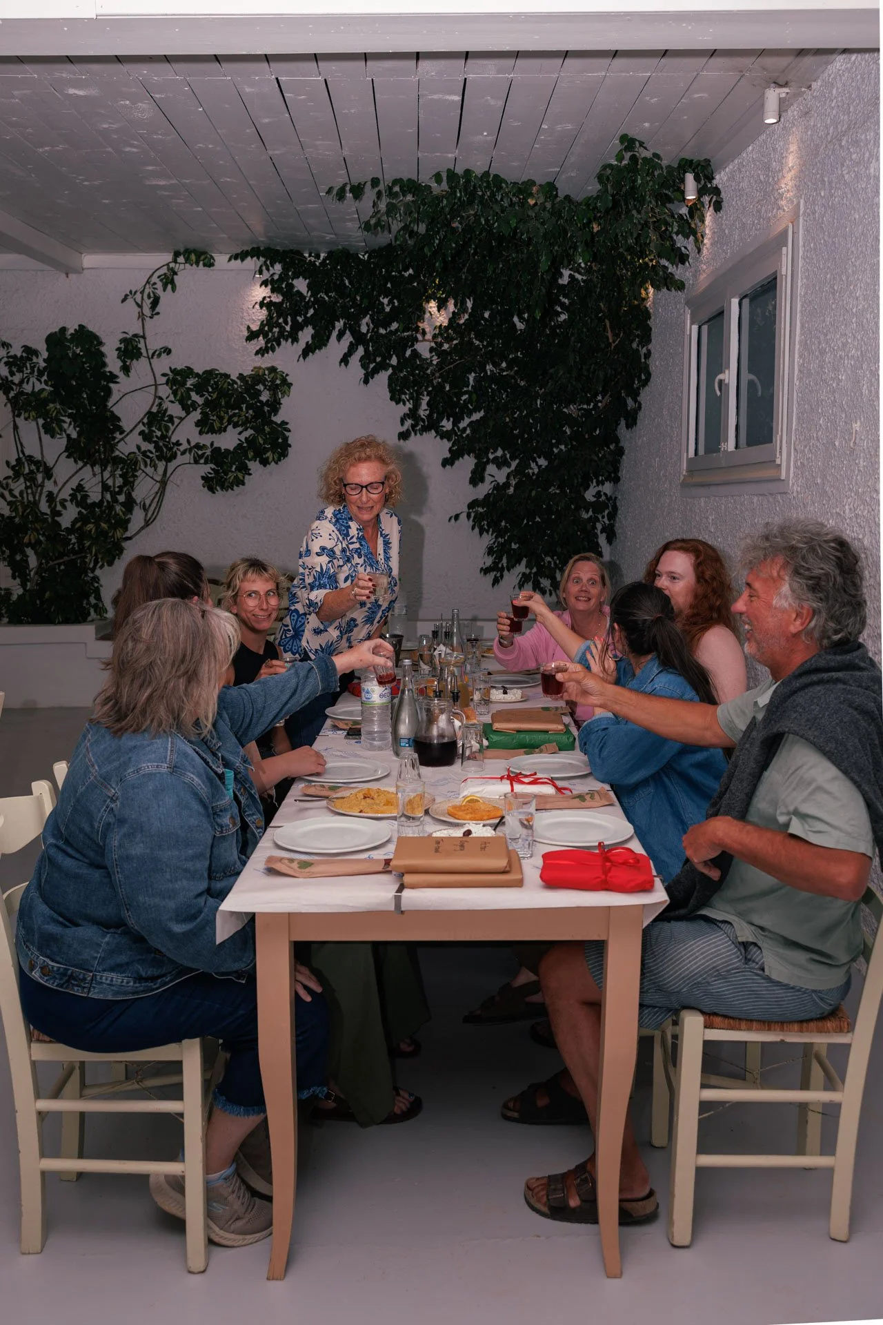 A group of people celebrating around a dining table, raising glasses for a toast, indoors with plants and white walls.