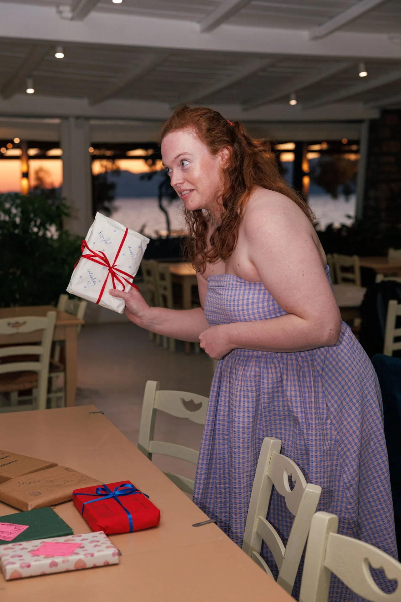 A woman with red hair in a purple and blue checkered dress holding a wrapped gift with a red ribbon, standing at a table with more wrapped presents, in a restaurant or cafe near a lake during sunset.