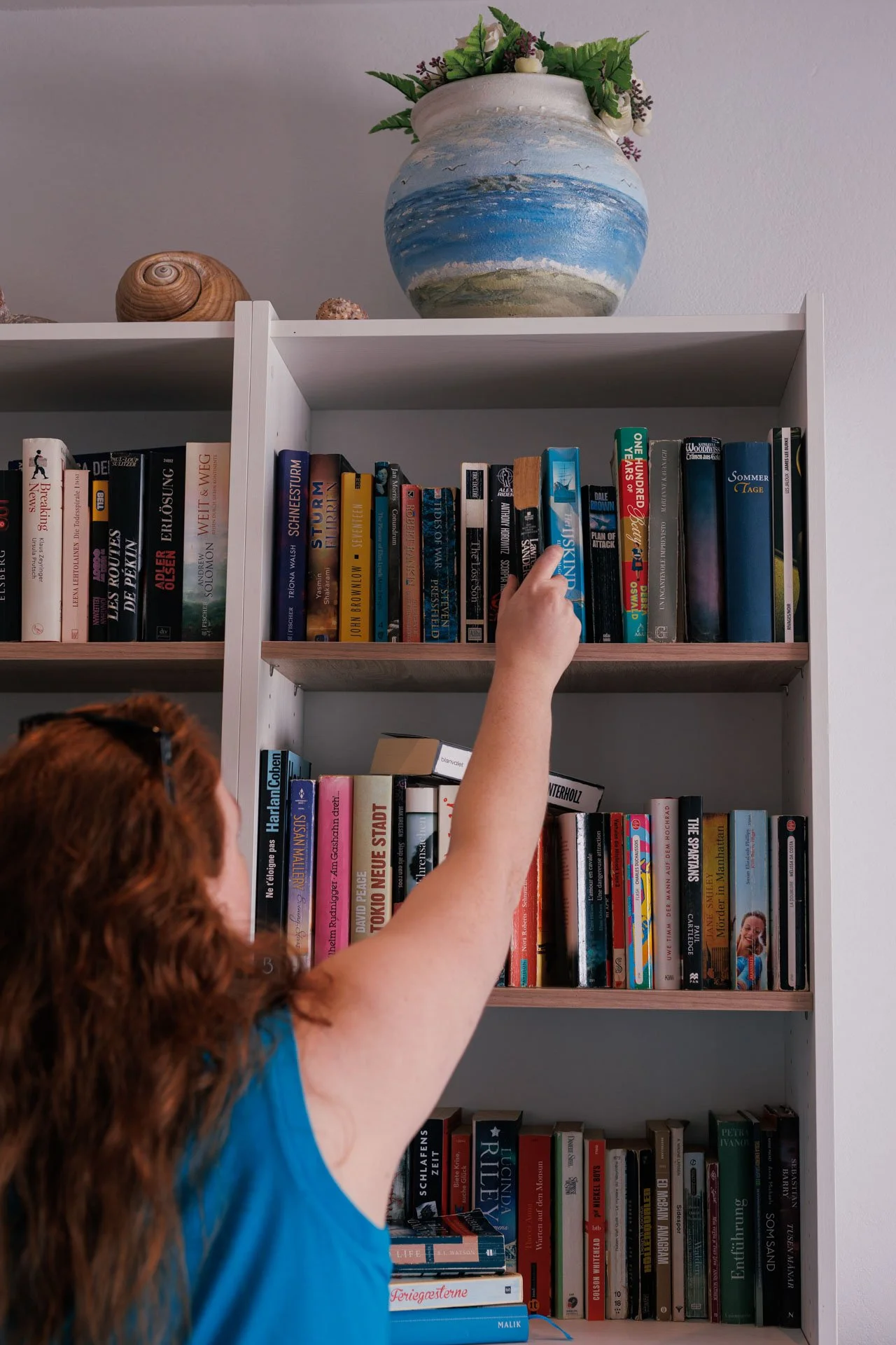 A person with red hair and a blue shirt reaching for a book on a multi-shelf white bookcase filled with various books. There is a large decorative vase with flowers on top of the shelf.