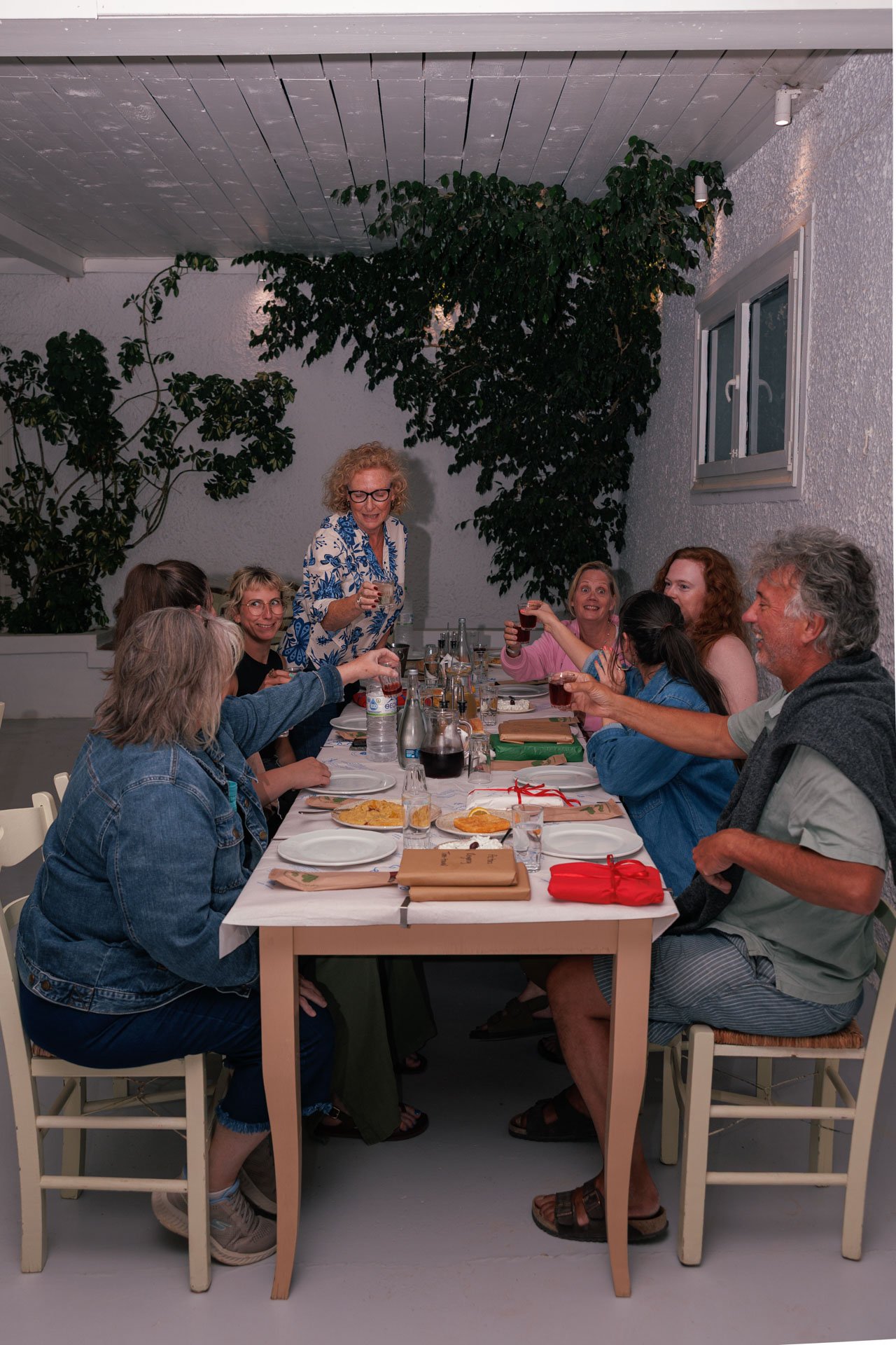 A group of eight people sitting around a dining table outdoors at night, raising glasses in a toast, with gifts and food on the table, under a white ceiling with planted greenery.