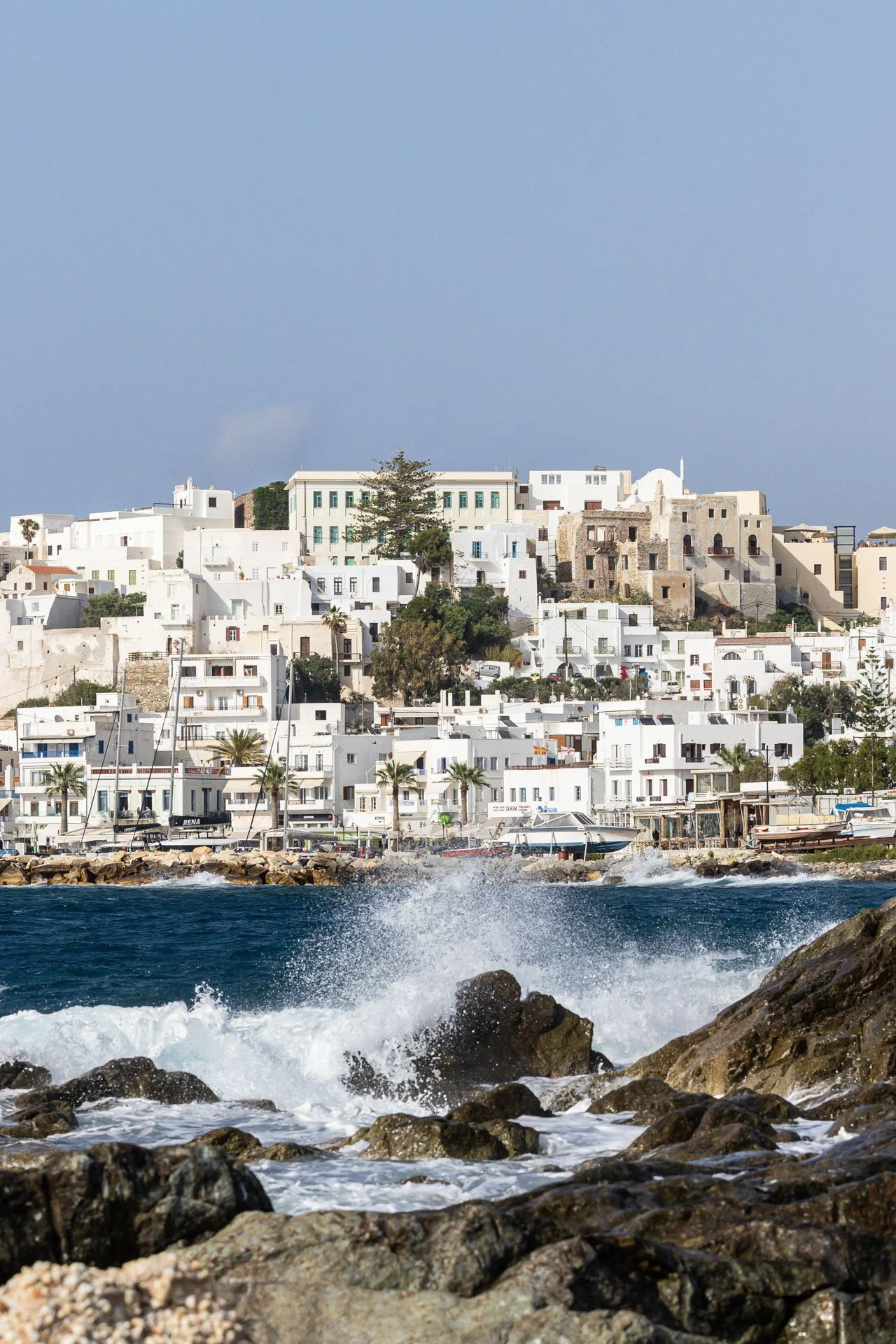 White buildings with flat roofs on a hillside along the coast, with rocks and waves crashing in the foreground, under a partly cloudy sky.