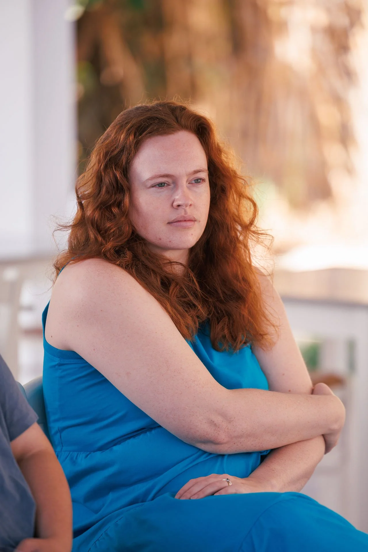 A woman with red curly hair wearing a blue sleeveless top and pants, sitting outdoors with her arms crossed and a serious expression.