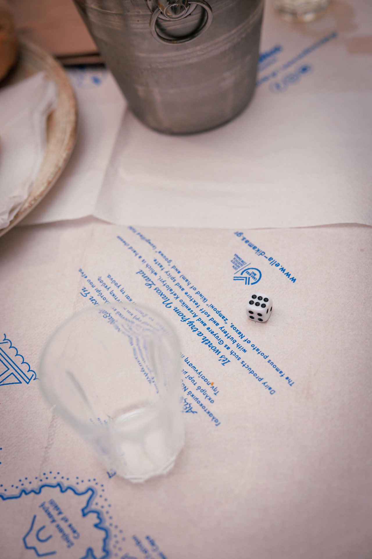 A tabletop with a clear glass tumbler, a white paper napkin with blue printed text and illustrations, a metal bucket, and a six-sided die showing the number five.