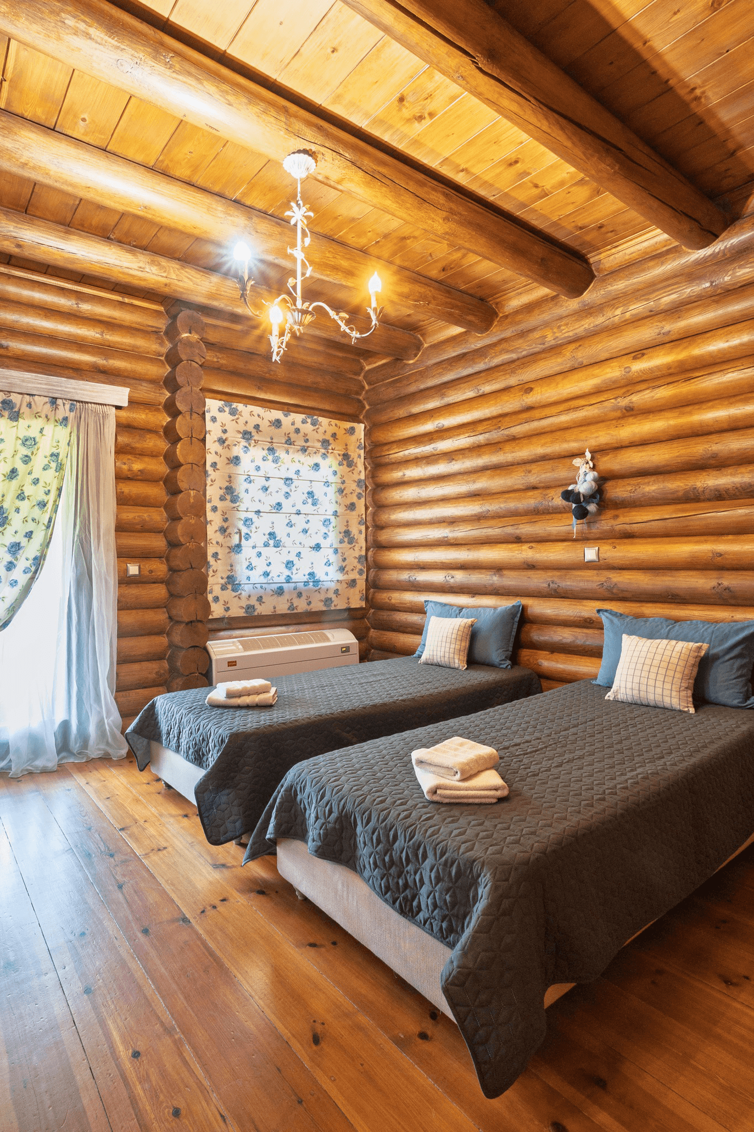 Two single beds in a log cabin bedroom with wooden walls and ceiling, each bed covered with a dark quilt, with folded towels on top, and a window with floral curtains.
