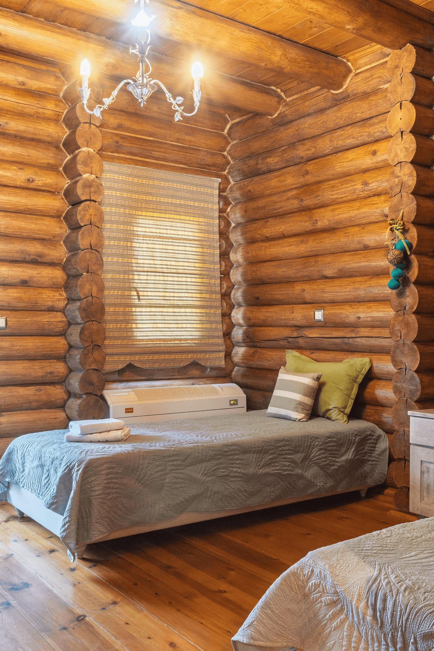 A bedroom in a log cabin featuring two single beds with graybedspreads and pillows, one with green and striped pillow, an air conditioner under a window with a striped curtain, and a wooden nightstand, illuminated by a chandelier with three bulbs.