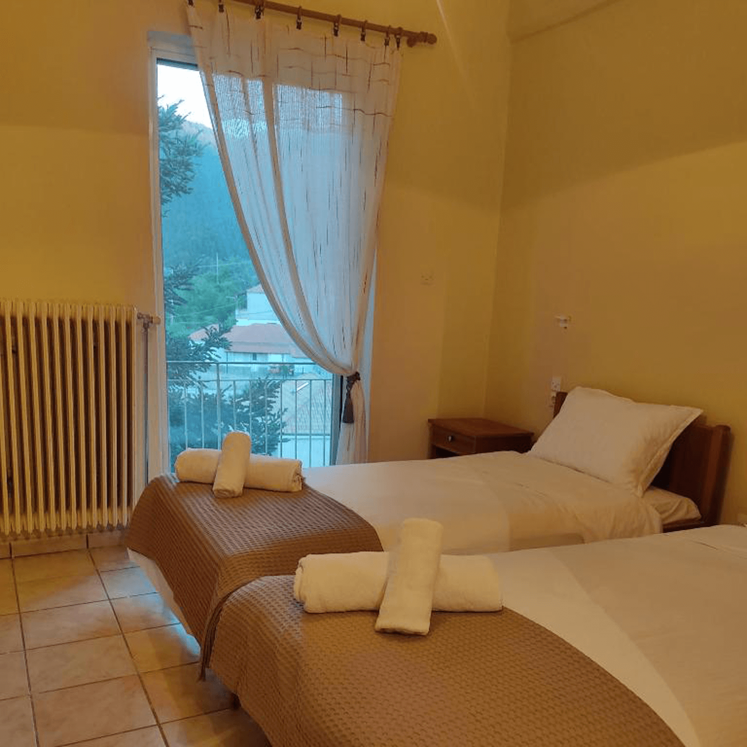 A hotel room with two single beds, each with white bedding and a pillow, and a small wooden nightstand in between. Towels are neatly rolled and placed at the foot of each bed. A window with sheer curtains lets in natural light and shows a view of tre