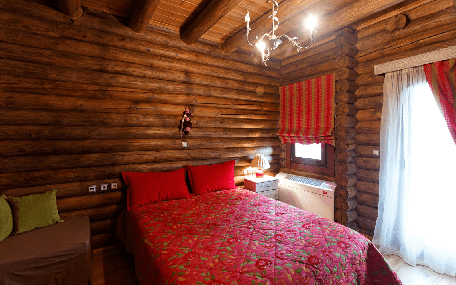 A cozy log cabin bedroom with wooden log walls and ceiling, a bed with red and green floral bedding, two red pillows, a nightstand with a lamp, a window with red and cream curtains, and a chandelier hanging from the ceiling.