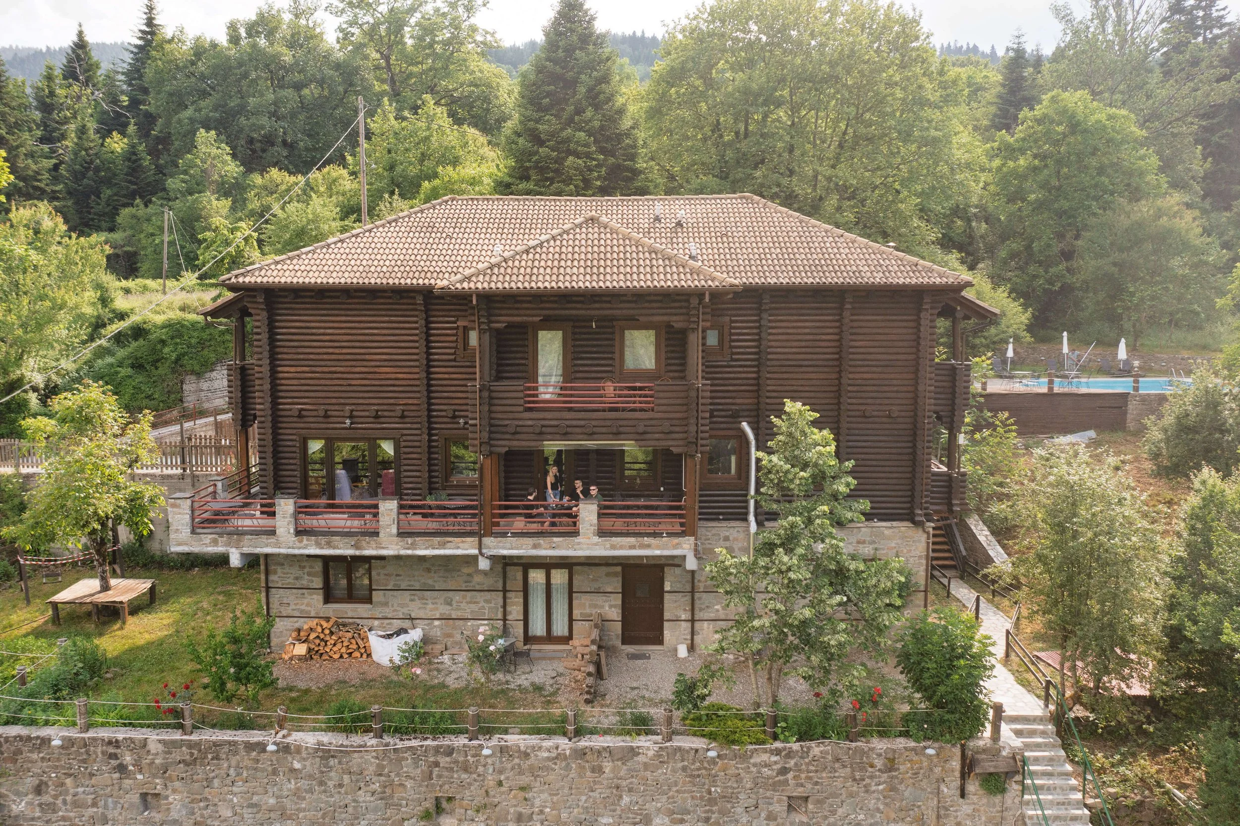 A large wooden log house with a tiled roof in a forested area, featuring a front porch with a railing, some people gathered on it, and a garden with trees and a stone wall in the yard, with a swimming pool and lounge chairs visible in the background.