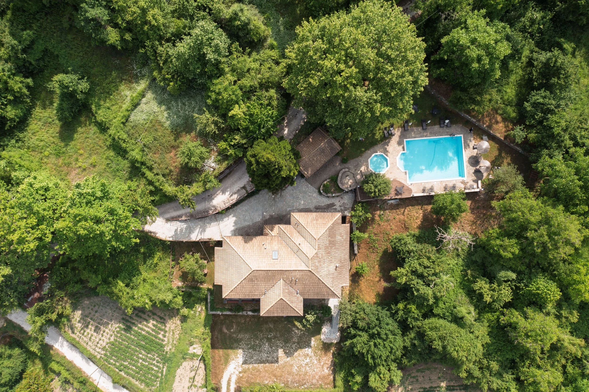 Aerial view of a house with a tiled roof, surrounded by trees, with a paved driveway leading to a swimming pool and a small hot tub on a patio, and a garden with crops nearby.