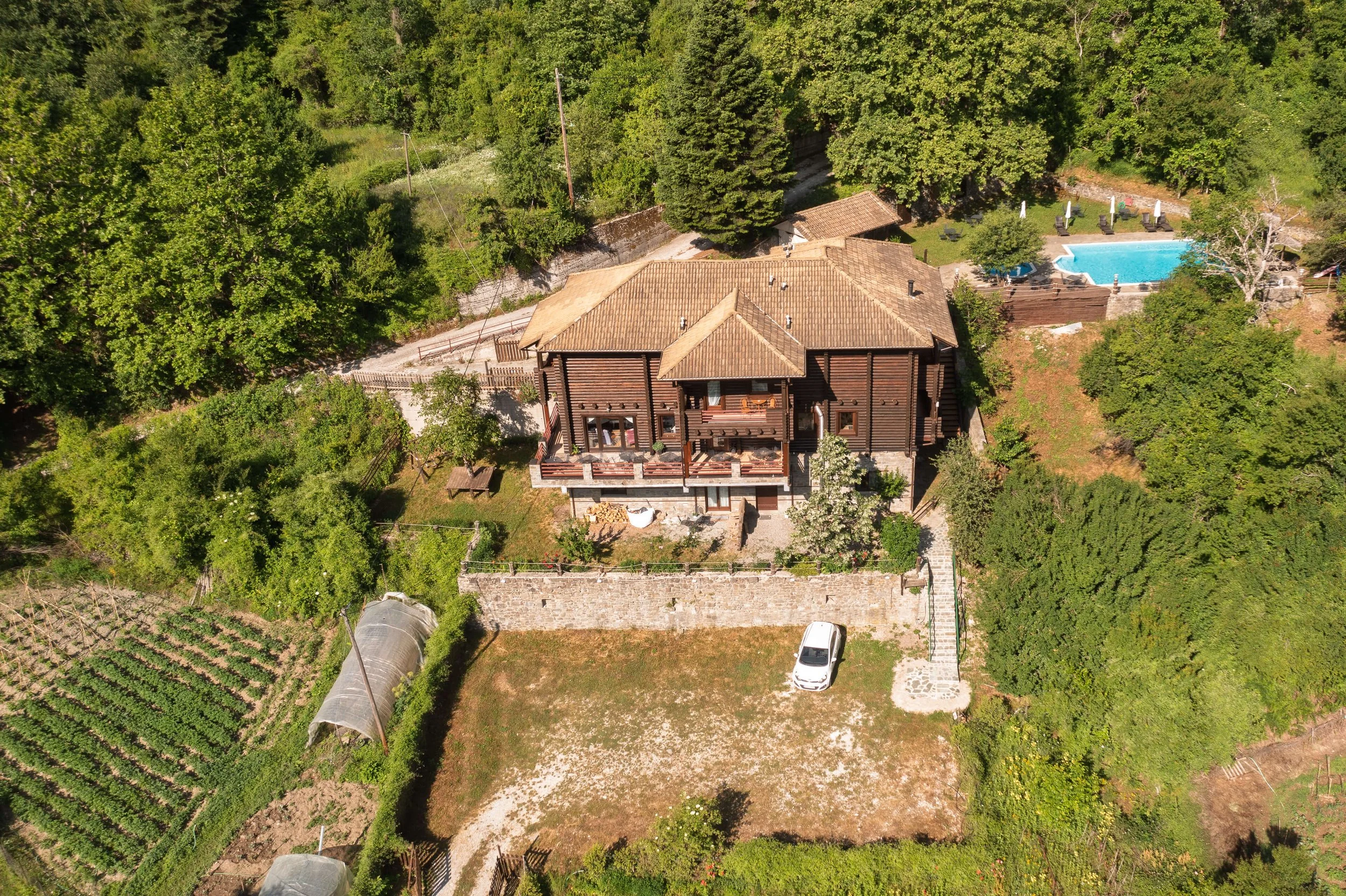 Aerial view of a large house with a brown tiled roof, surrounded by green trees, a garden, and a small pond with a pool nearby.