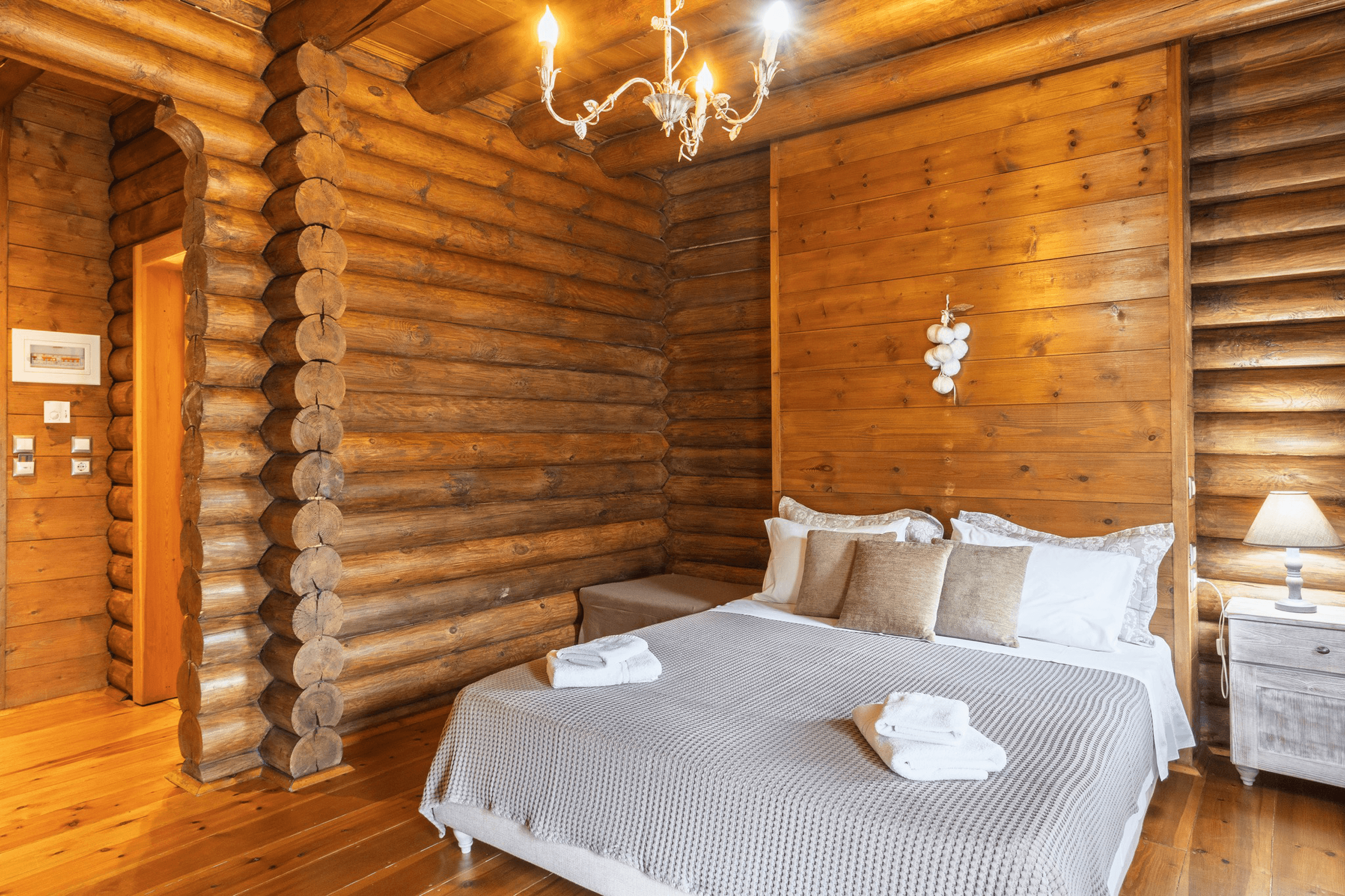 A cozy bedroom in a log cabin with wooden walls, a chandelier, a bed with white and beige linens, and a nightstand with a lamp.