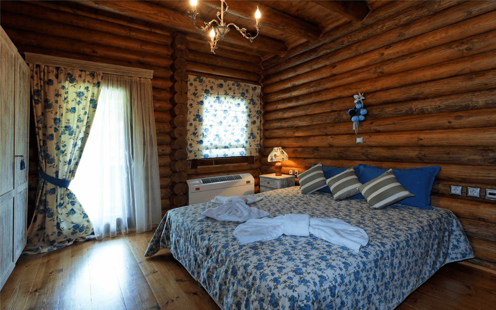 A cozy log cabin bedroom with a bed decorated with a floral bedspread and striped pillows. There is a window with floral curtains, a small nightstand with a lamp, and wooden log walls and ceiling. Towels are folded on the bed.