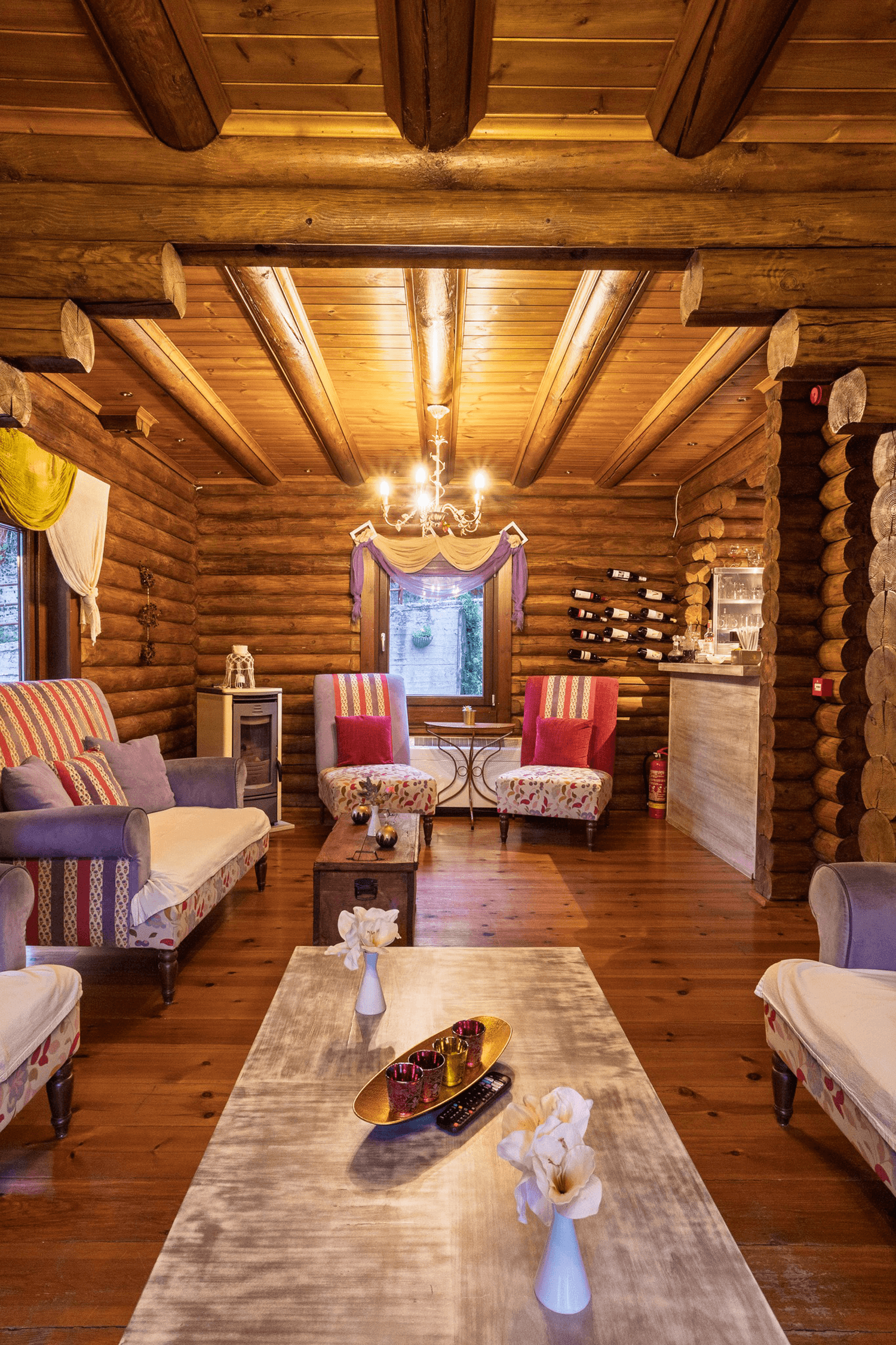 A cozy living room with wooden log walls and ceiling, featuring a chandelier, two armchairs with red cushions, a sofa with patterned fabric, a wooden coffee table with decorative items, and a bar area with wine bottles and glasses.