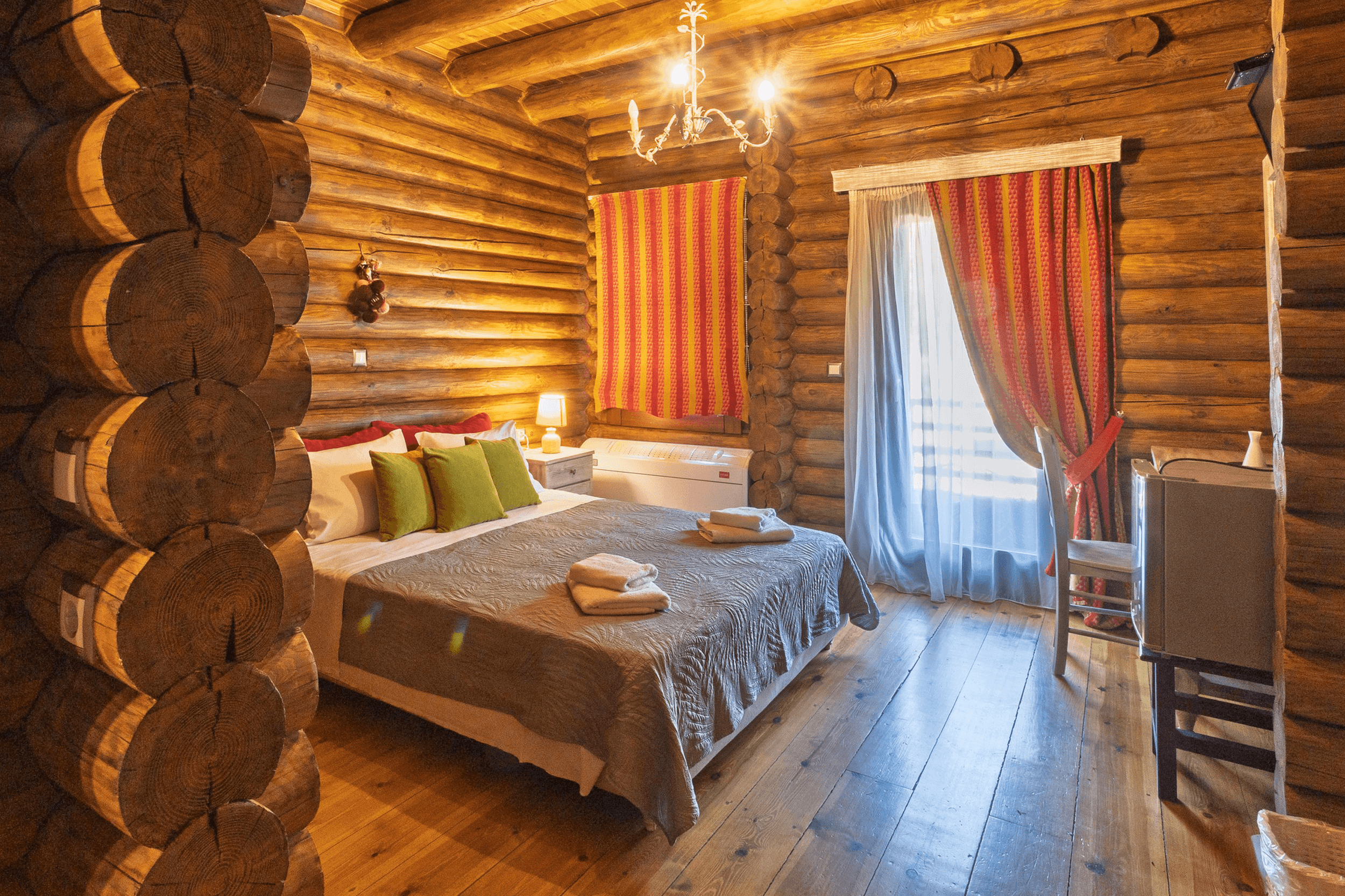 A rustic log cabin bedroom with a large bed, green pillows, a bedside table with a lamp, red and yellow striped window coverings, a chandelier, and hardwood floors.