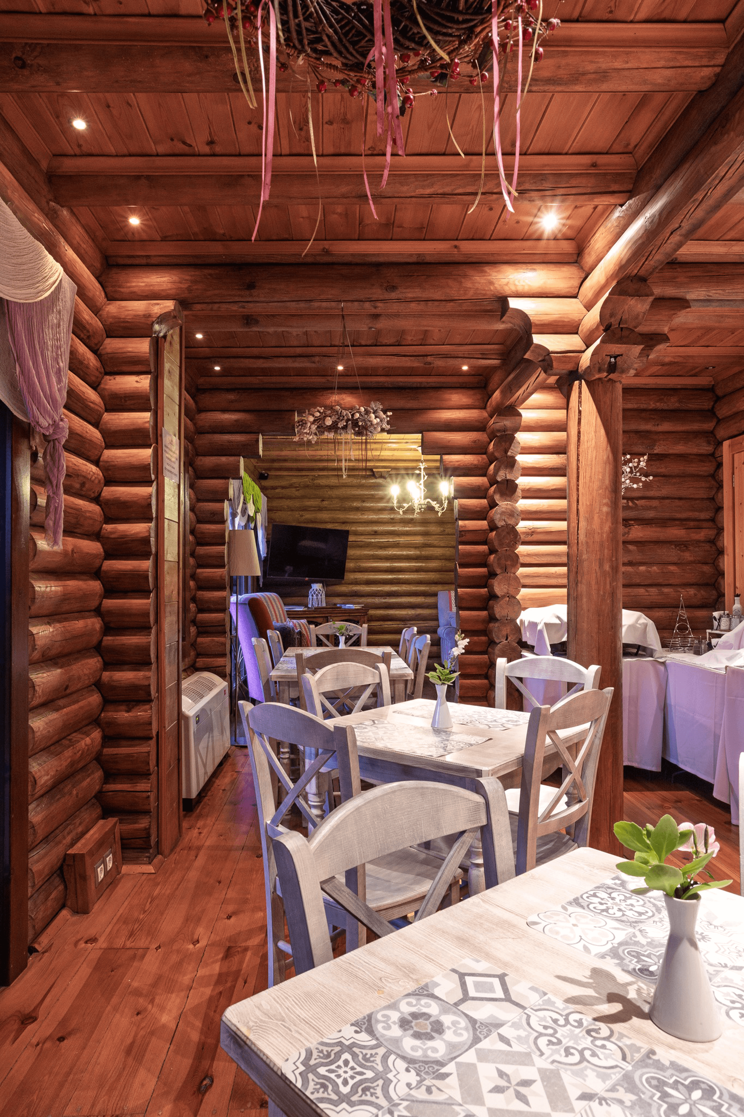 Interior of a cozy log cabin restaurant with wooden walls and ceiling, decorated with hanging ribbons and flowers, white tables with lace tablecloths, potted plants, and a mix of chairs. Includes a chandelier and a TV in the background.