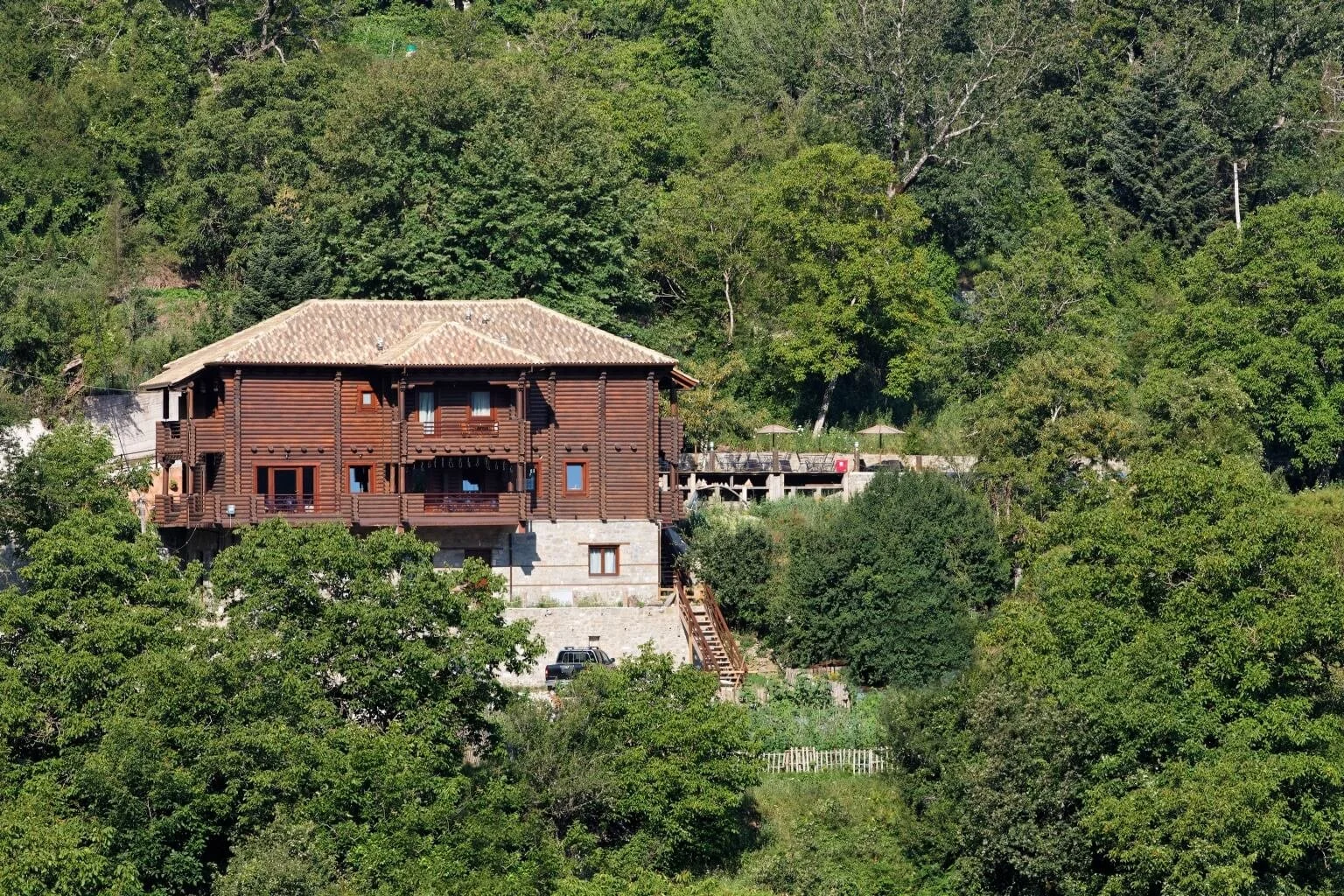 A large wooden house on a hillside surrounded by dense green trees.