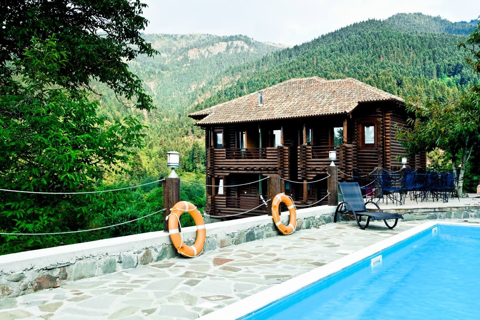 A scenic mountain landscape with a wooden cabin, outdoor pool, poolside lounge chairs, and patio furniture, surrounded by lush greenery and forested hills in the background.